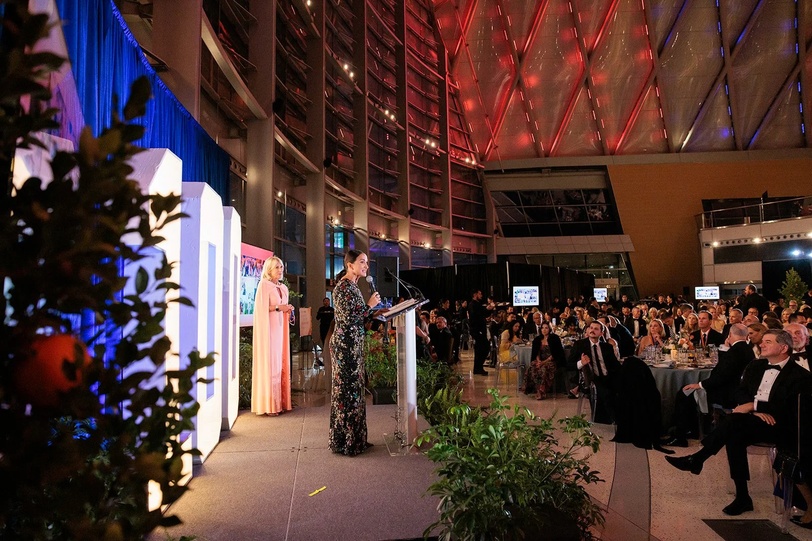 A woman giving a speech at a formal event in a large, modern conference hall with a high, colorful ceiling. Many guests are seated at round tables, listening attentively, dressed in formal attire.