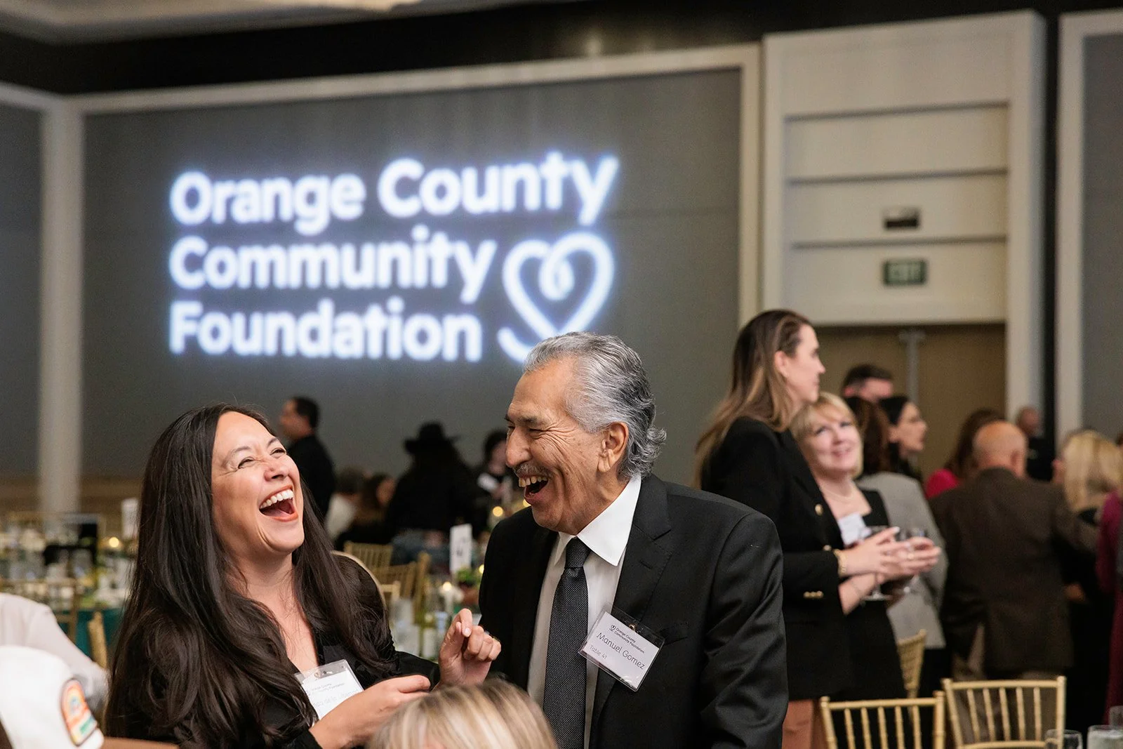 Two people laughing and talking at a formal event, with a large sign in the background reading 'Orange County Community Foundation'