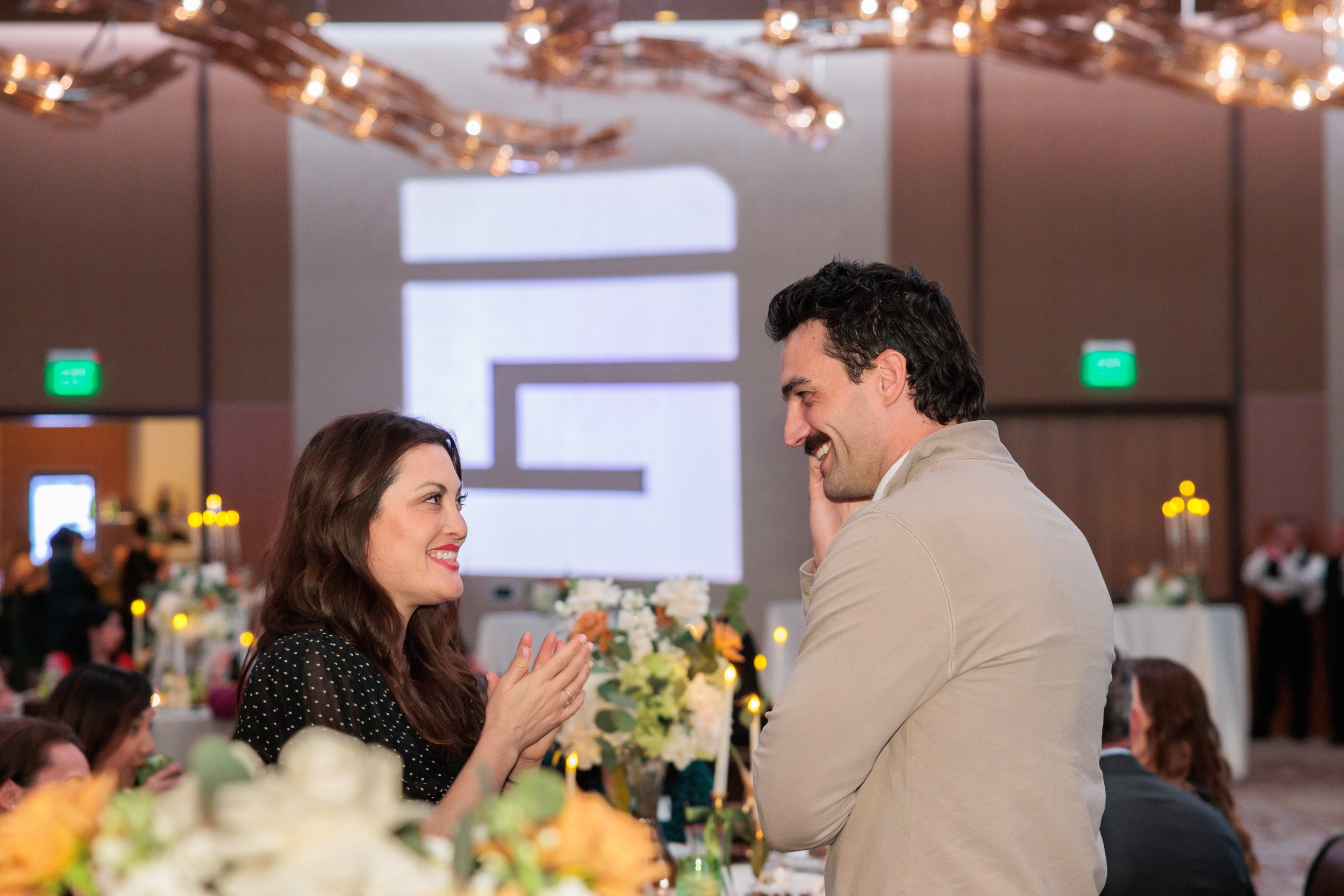 A woman and a man are smiling and gazing at each other at a formal event, with a decorated table and a patterned backdrop in the background.