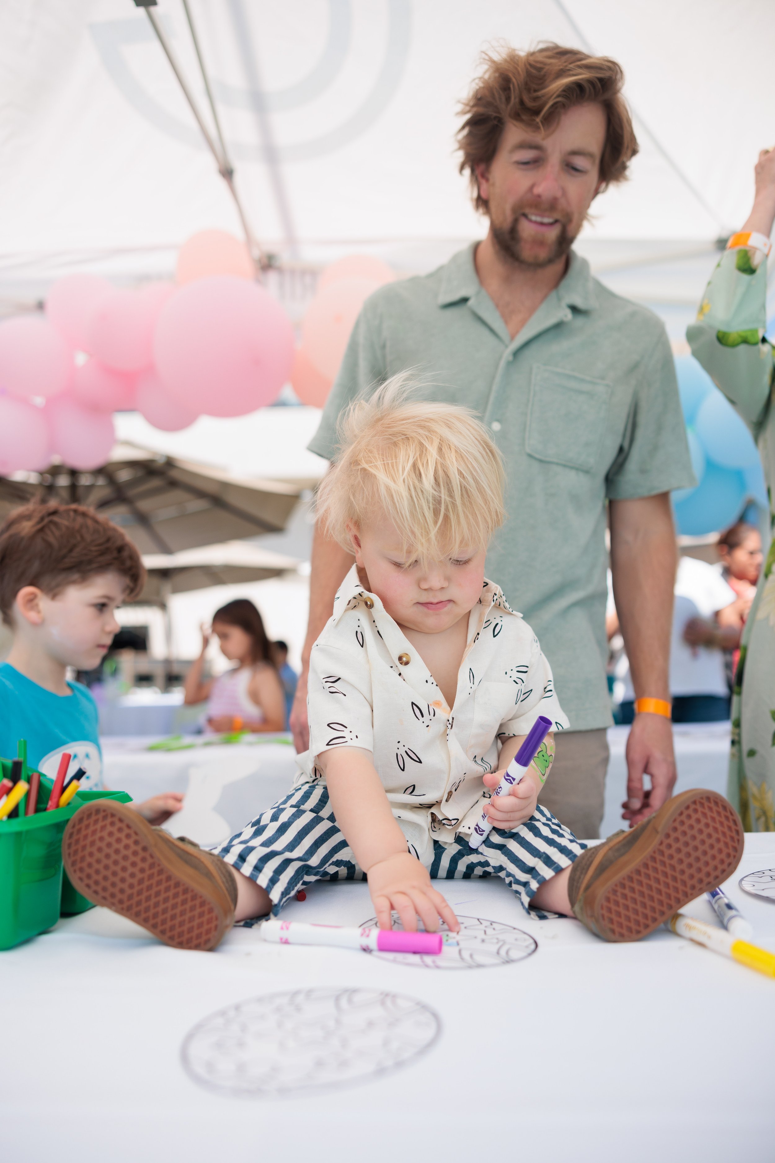 A young blonde boy sitting on a table, drawing with colorful markers, with an adult man standing behind him. Another boy is nearby, and there are pink balloons and outdoor umbrellas in the background.