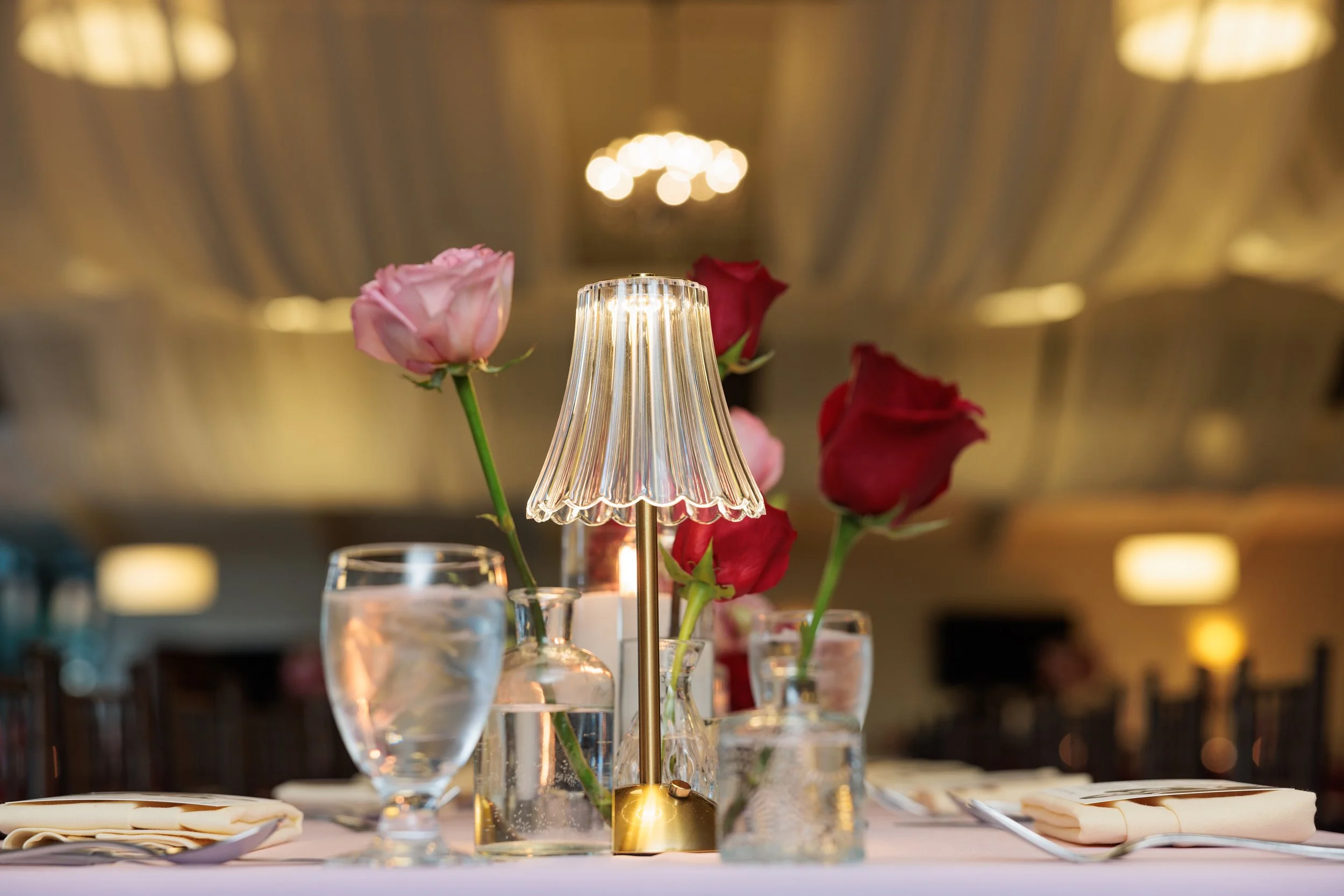 Elegant table centerpiece with pink and red roses in glass vases, a small brass lamp, and water glasses, set in a warmly lit banquet hall.