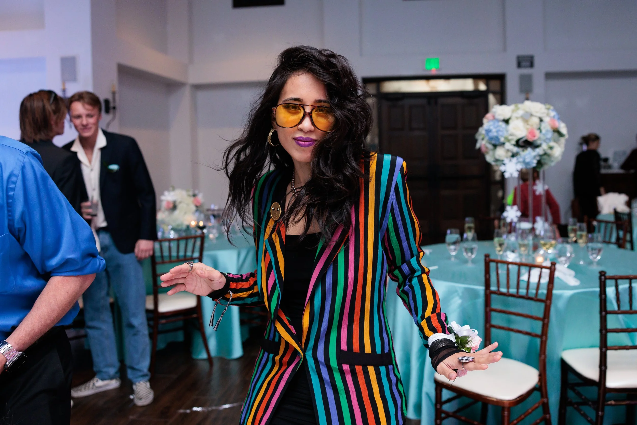 A woman with long dark hair wearing yellow-tinted sunglasses and a colorful striped blazer, standing at a decorated event with tables and floral centerpieces.