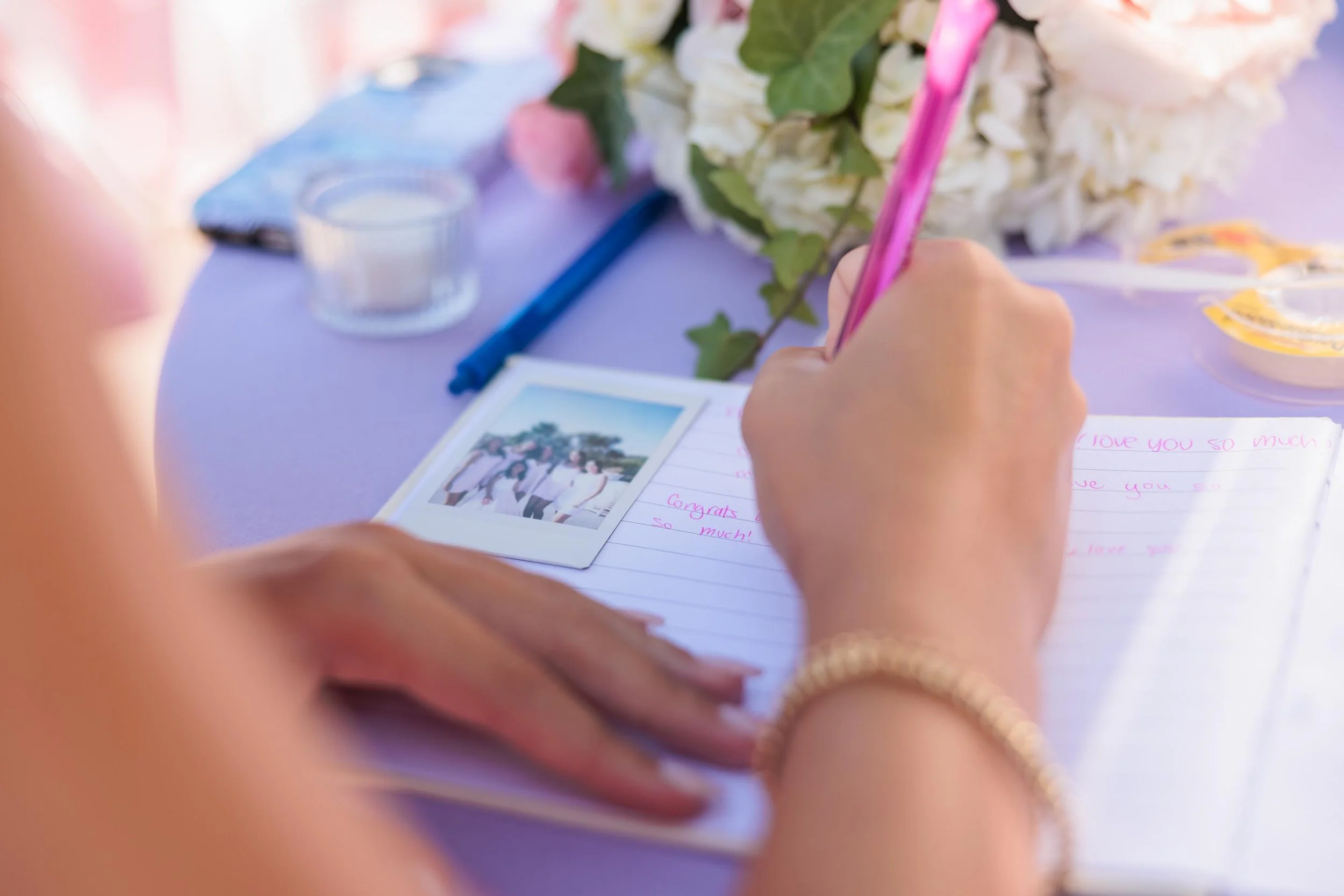A person writing in a notebook with a pink pen at a table decorated with flowers, a photograph, a glass of water, and other small items.