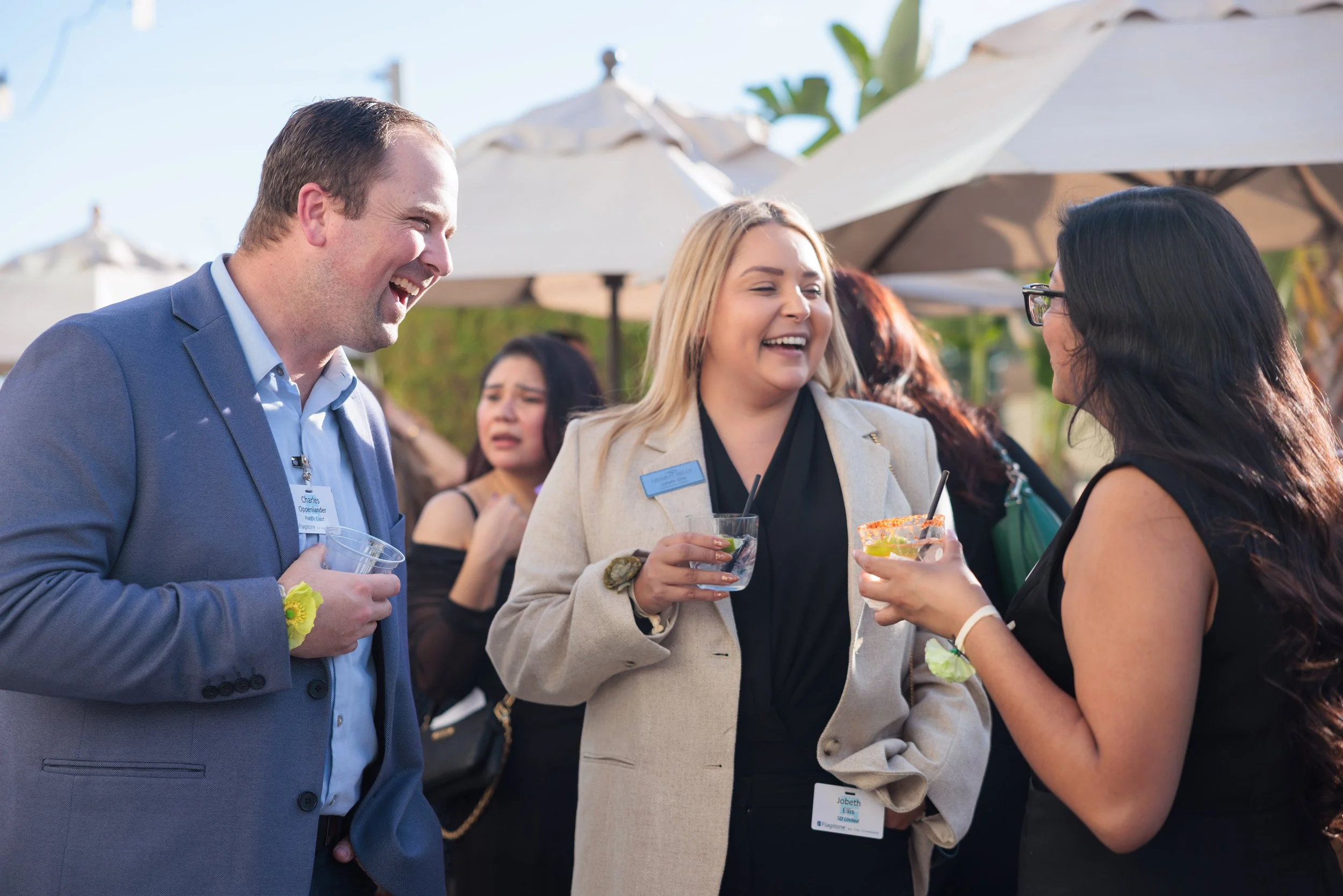 Group of people, two women and a man, at an outdoor event, talking and holding drinks, with umbrellas and greenery in the background.