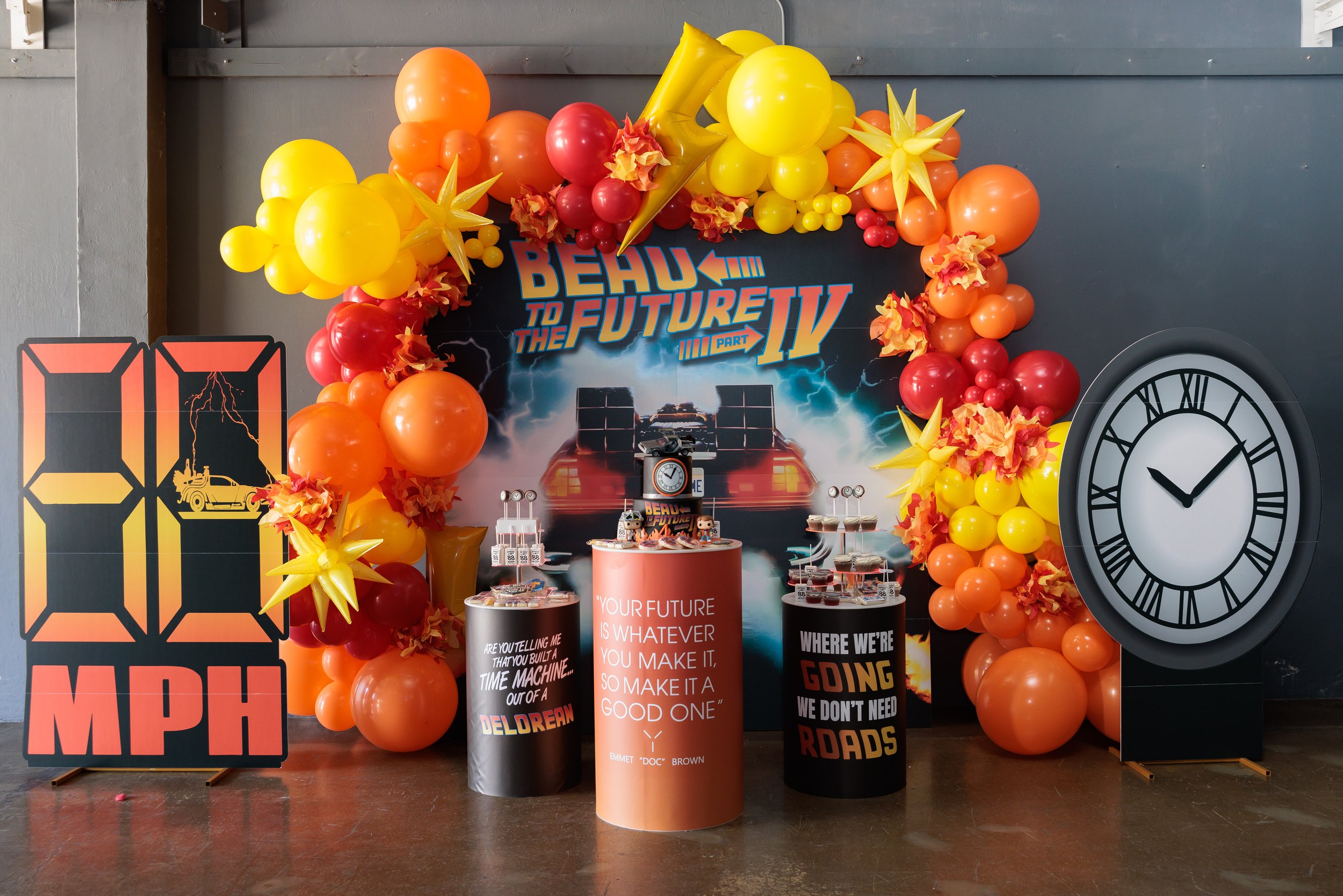 Decorated backdrop for 'Back to the Future IV' party featuring a graphic of a DeLorean, surrounded by orange, yellow, and red balloons and paper flowers, with two quote banners and a large clock to the right.