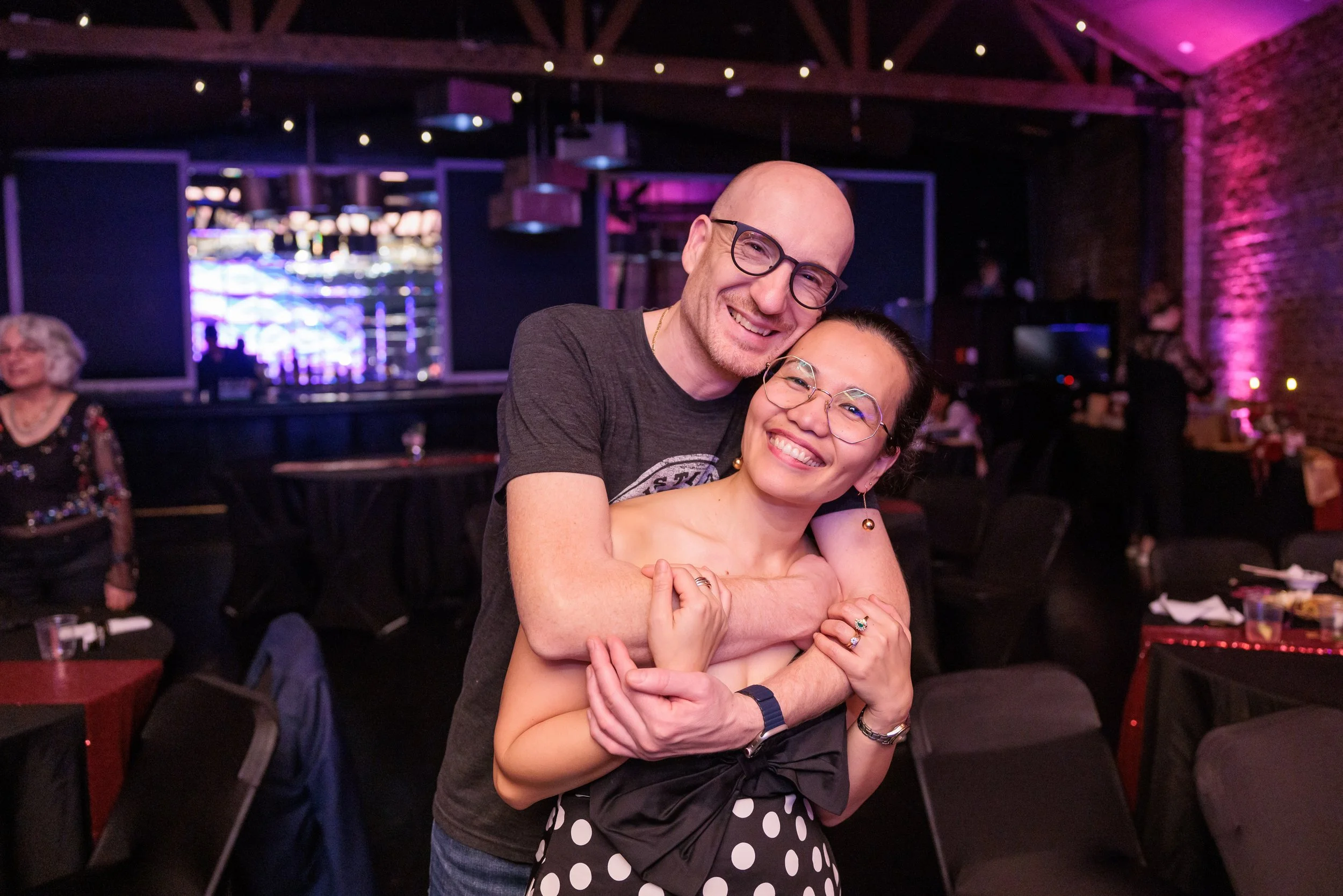 Two people hugging and smiling in a lively, decorated indoor setting, possibly a restaurant or event venue, with other people and tables in the background.