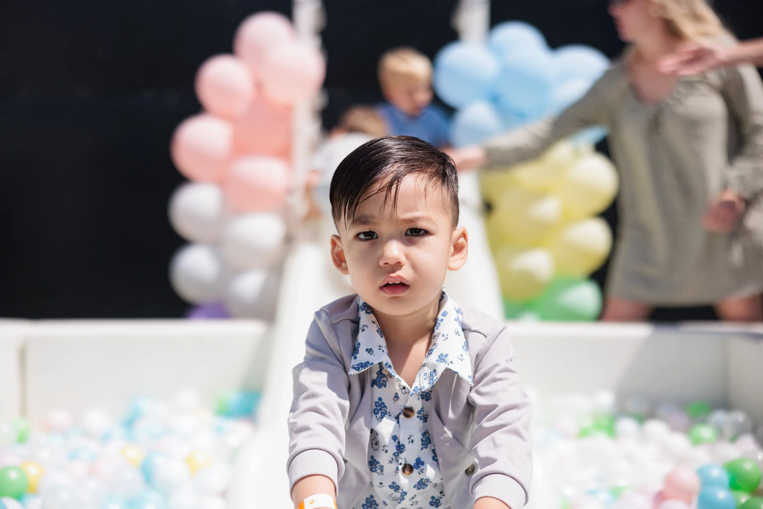 A young boy with dark hair and a patterned shirt looks confused in a ball pit at a playground, with blurred children and colorful balloons in the background.