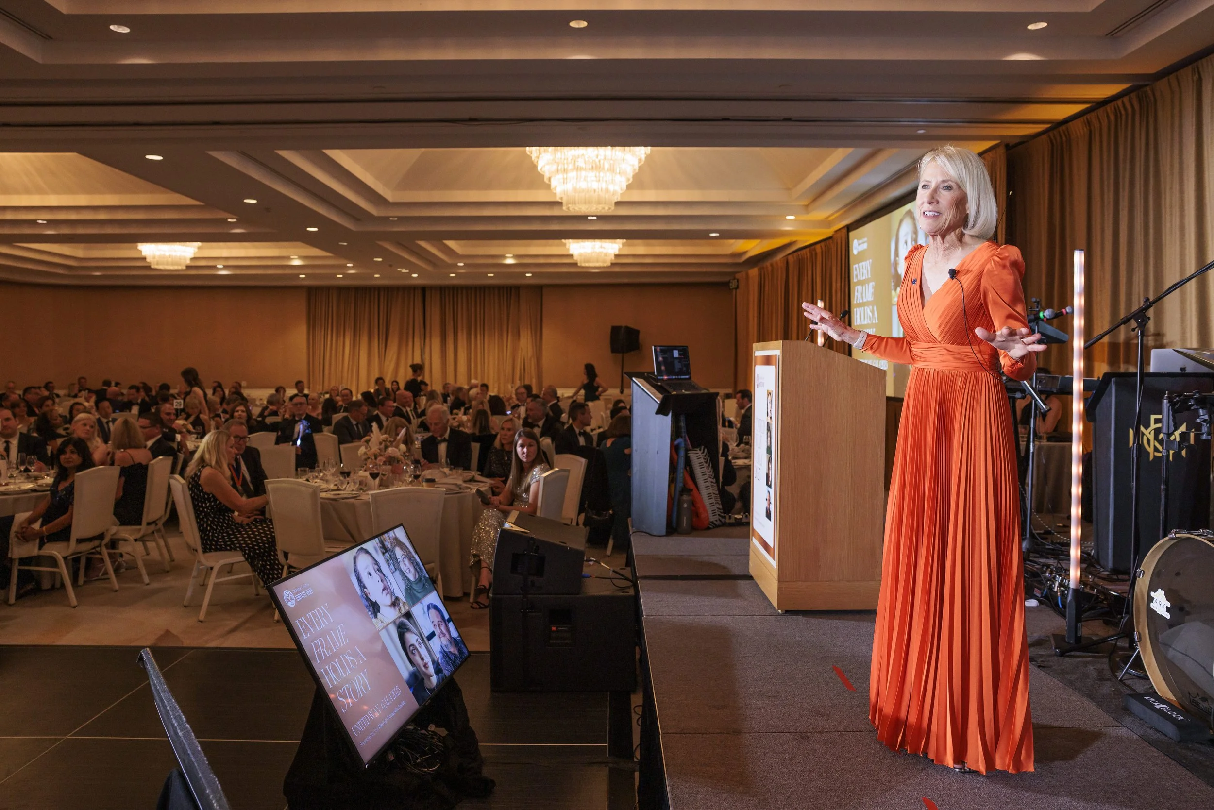 A woman in an orange pleated dress is speaking at a podium on stage in a large banquet hall with attendees seated at banquet tables.