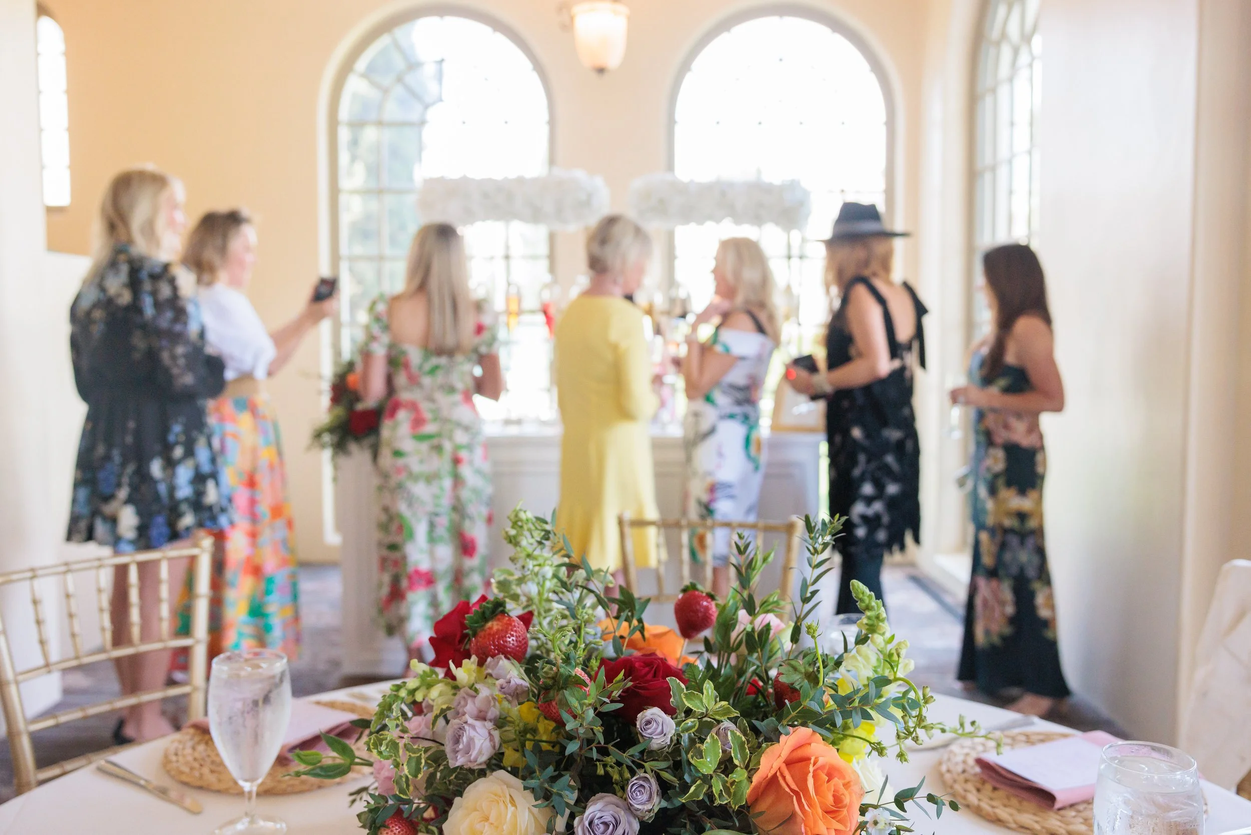 A group of women in colorful dresses are gathered in a bright room with arched windows, some holding flowers or glasses, and one woman is taking a photo with her phone. In the foreground, there is a table with a large floral centerpiece, water glasse