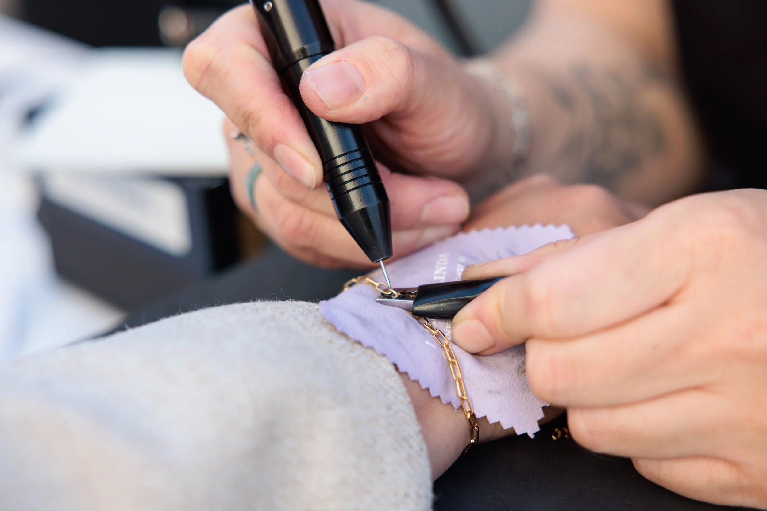 A close-up of a person's hands jewelry designing or repairing a gold chain using a handheld tool, with a jewelry cleaning cloth nearby.