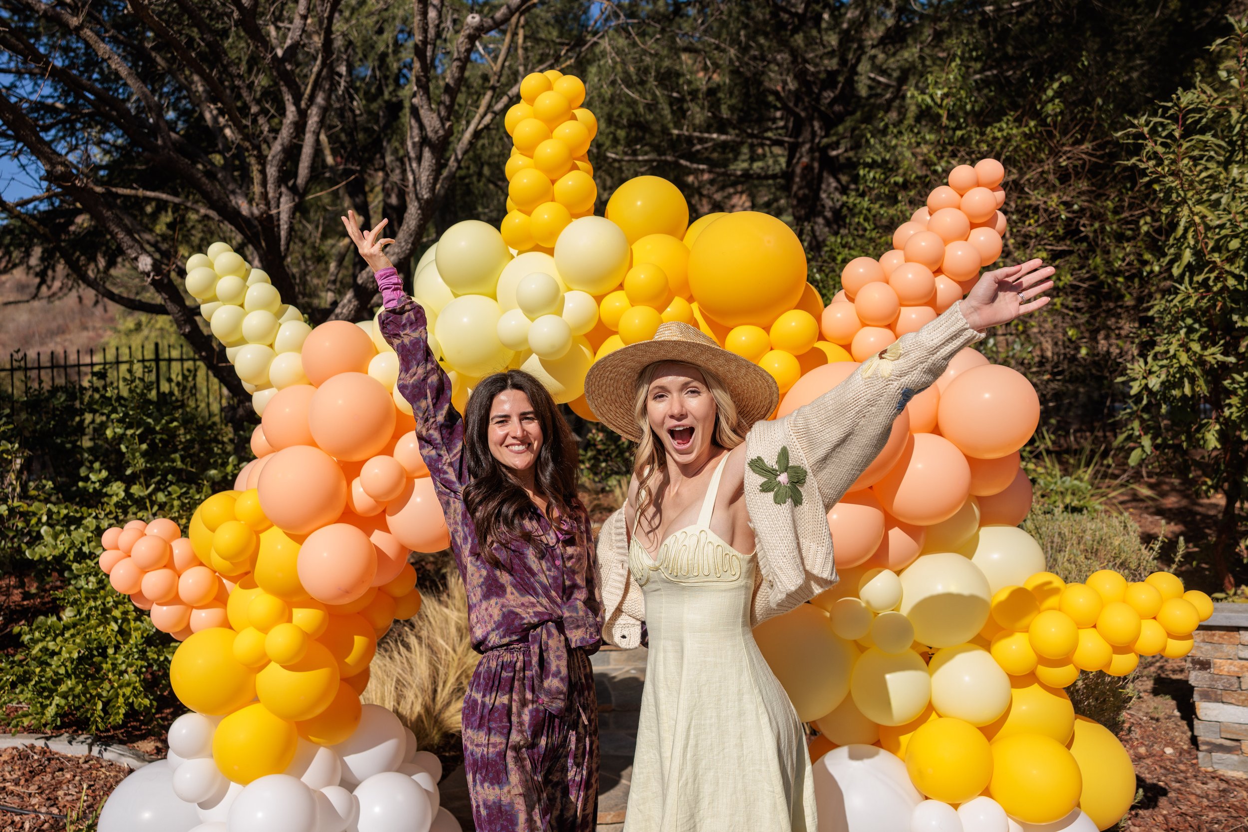Two women smiling and celebrating outdoors in front of a large decorative balloon arch with yellow, peach, and white balloons, surrounded by trees and greenery.