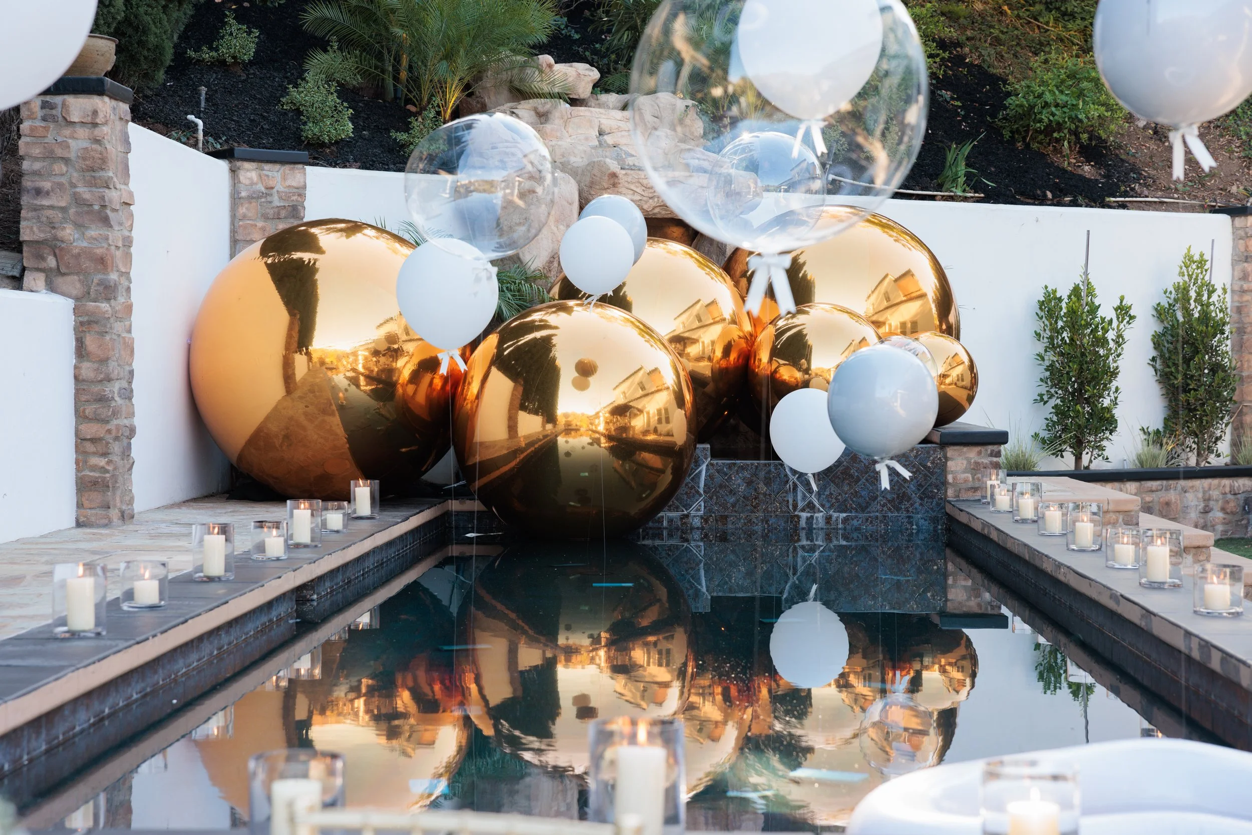 Decorative outdoor scene with large gold and white balloons, candles in glass holders lining a pool, and green shrubbery. The setting looks festive, possibly for a celebration or party.