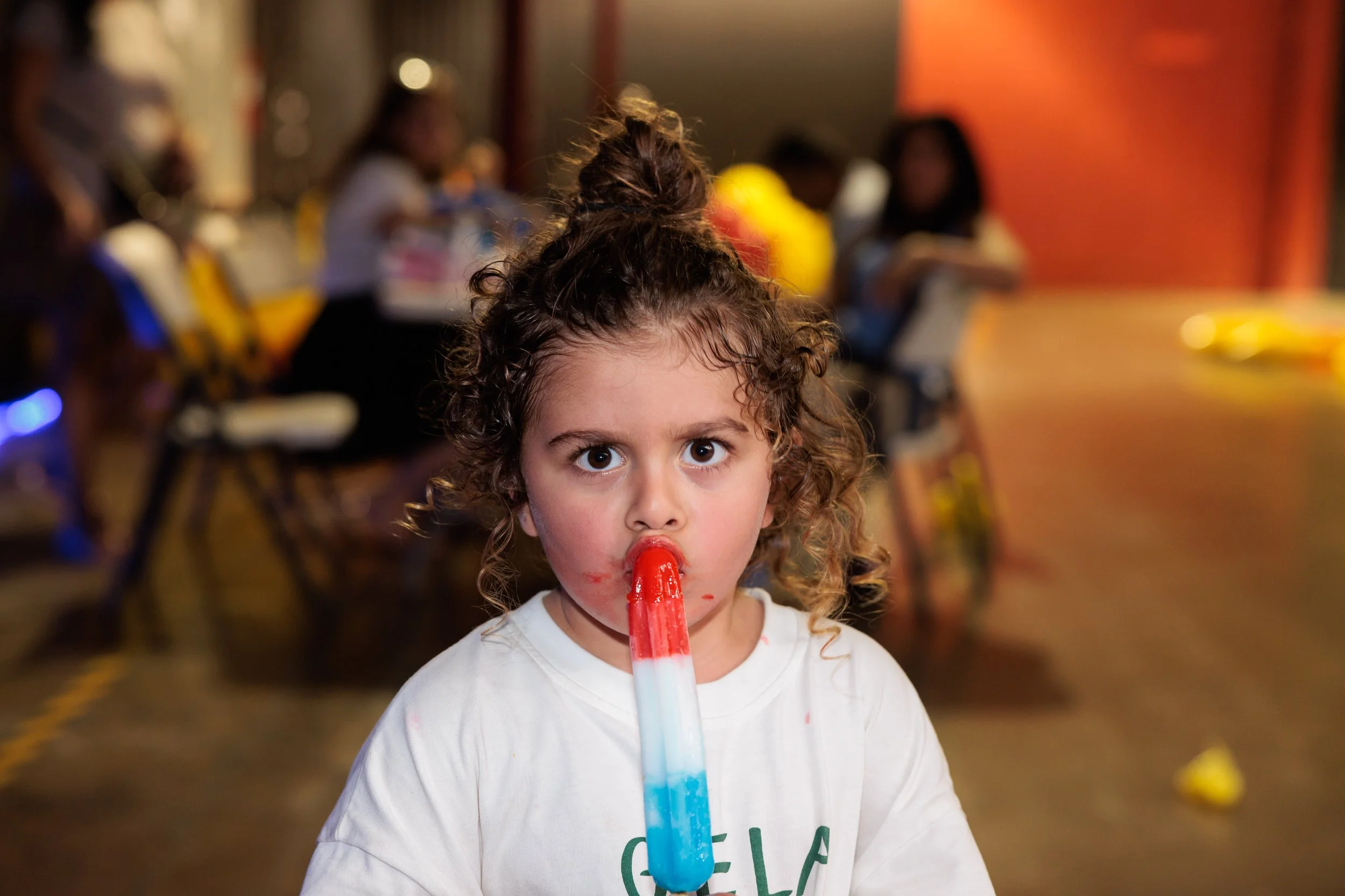 Young girl with curly hair and large eyes looking into camera while sucking on a red, white, and blue popsicle, with a blurry background of people sitting at a table indoors.