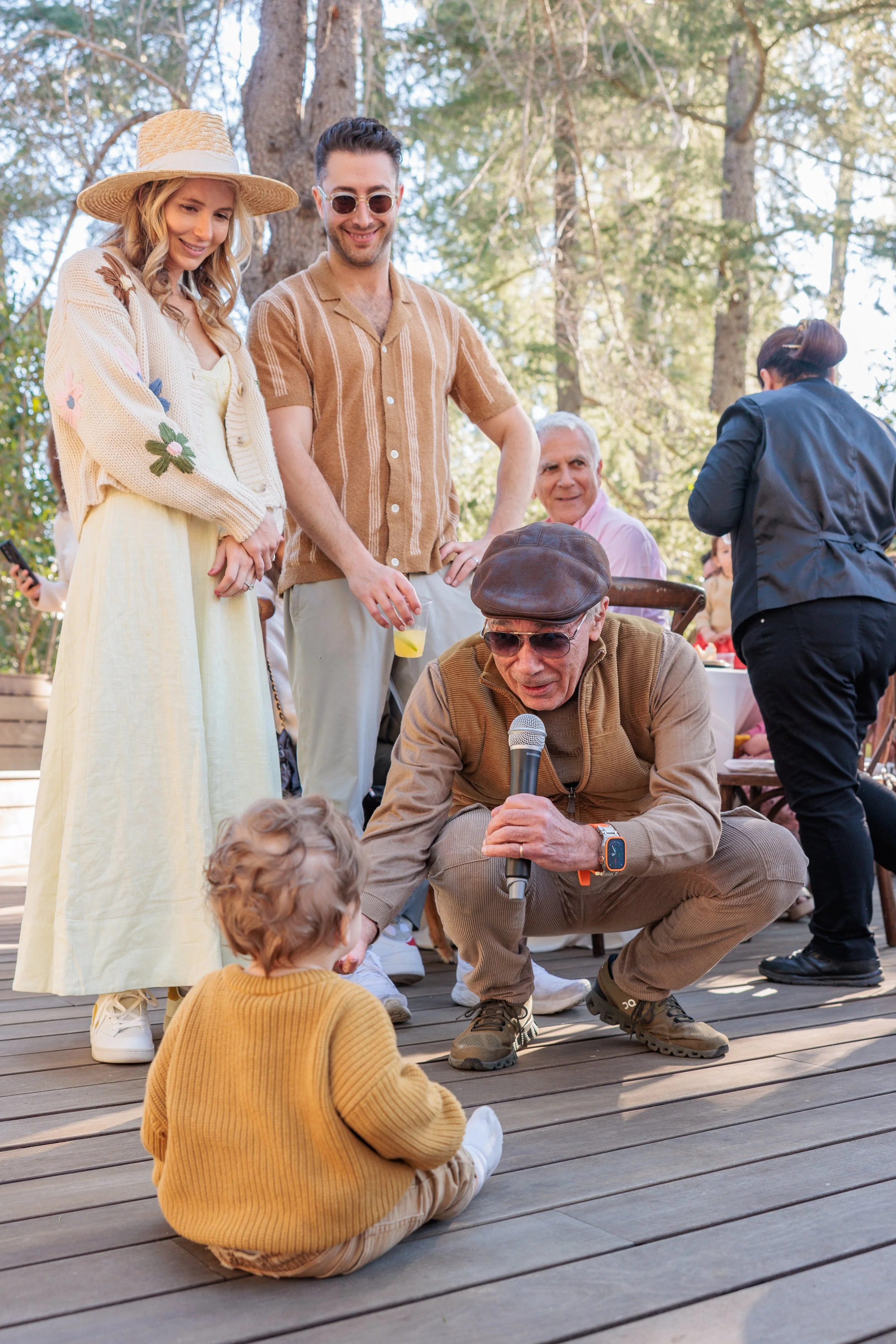 An elderly man is crouching down, holding a microphone and speaking to a young child sitting on a wooden deck, surrounded by a group of people outdoors in a forested area.