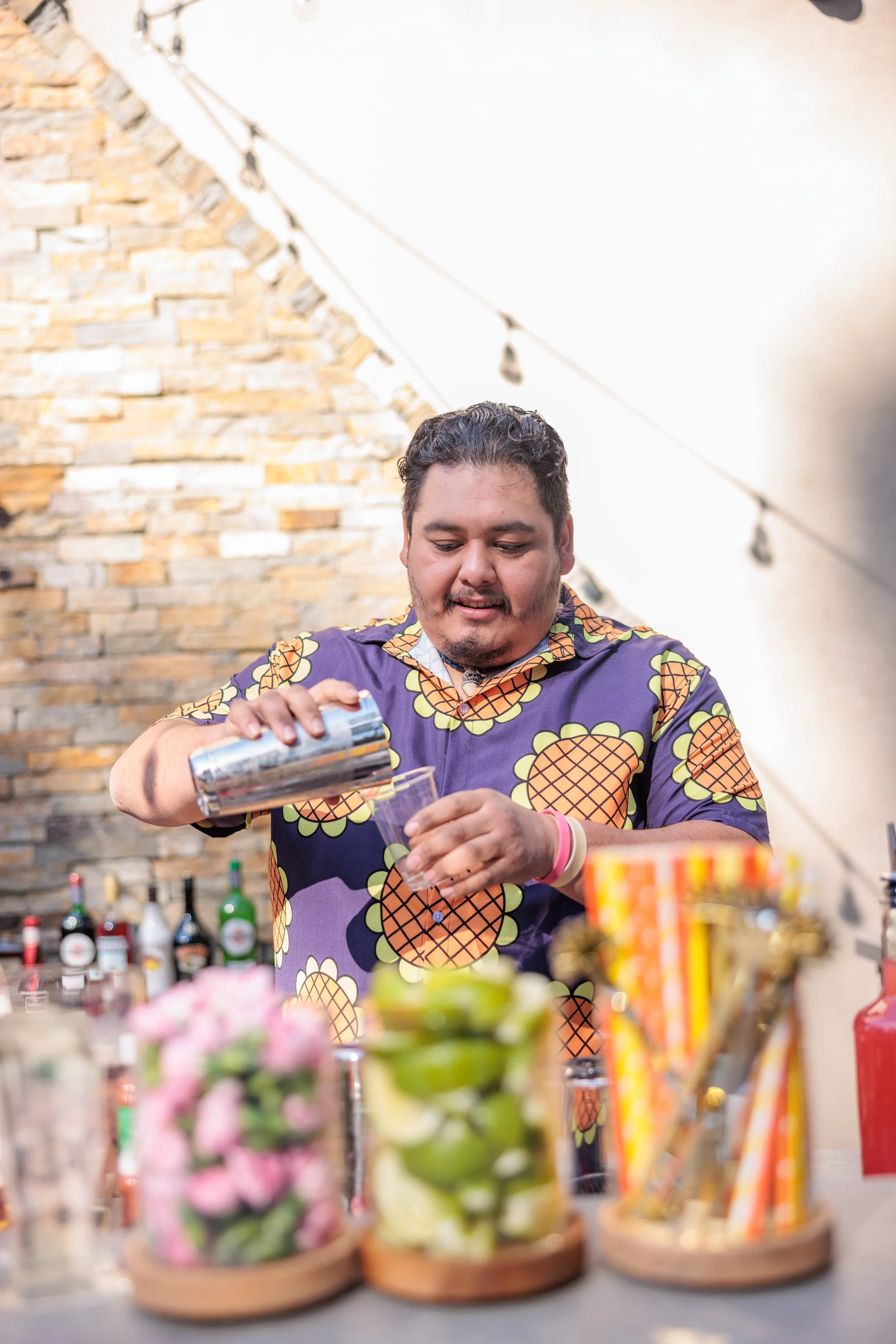 A man preparing drinks behind a bar with a brick wall background, bottles of alcohol, flowers, and straws on the counter.