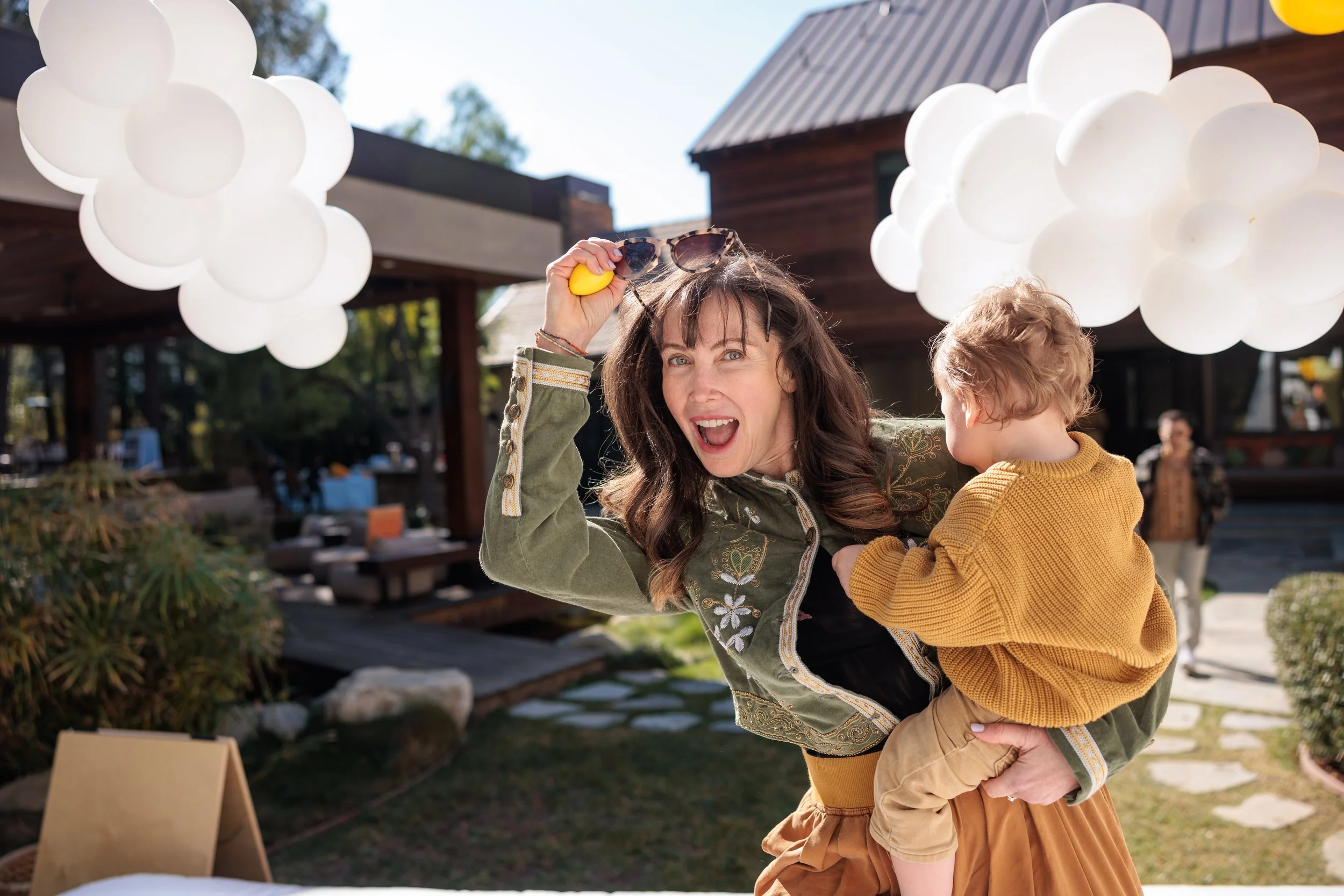 Woman holding a child outdoors during daytime, with balloon decorations in the background.