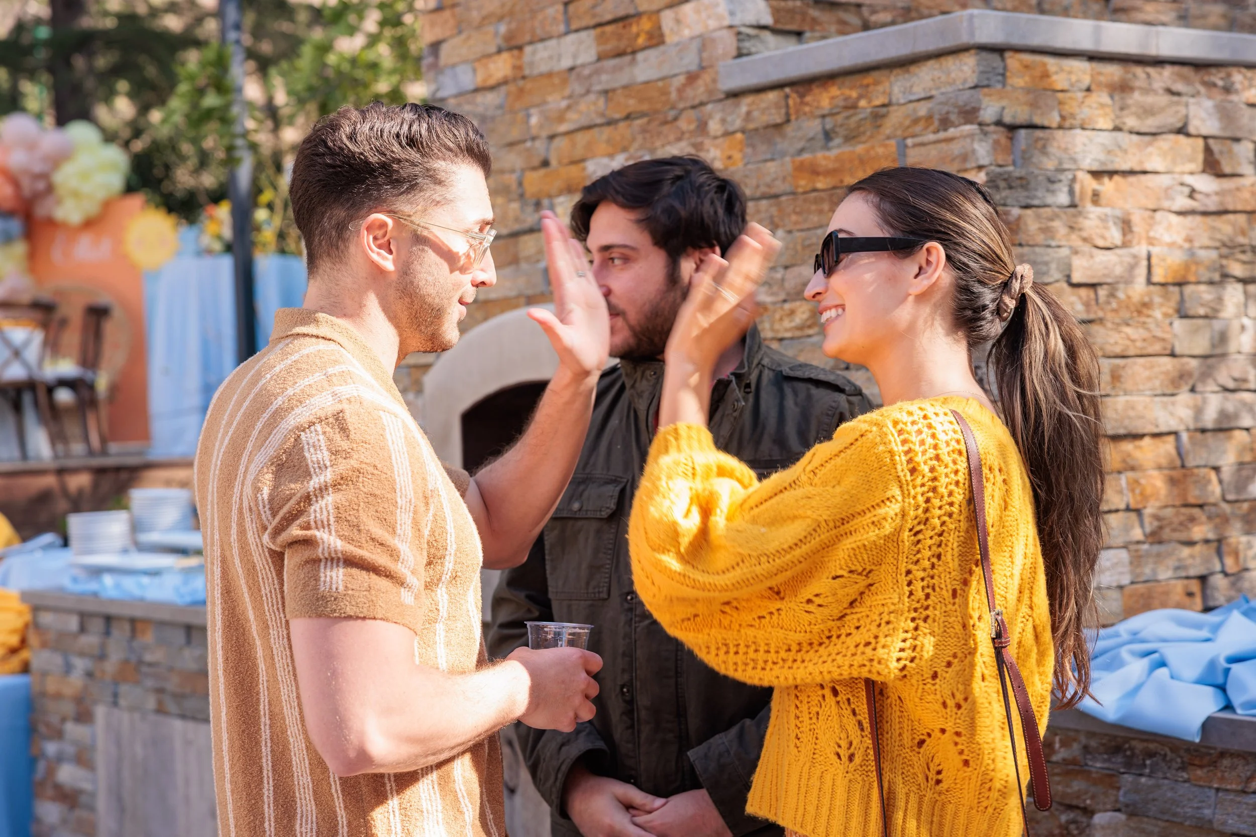Three people cheer and high-five outdoors during a sunny day, with a brick wall and outdoor furniture in the background. The woman wears sunglasses and a mustard yellow sweater, the man on the left has short hair and glasses, and the man in the middl