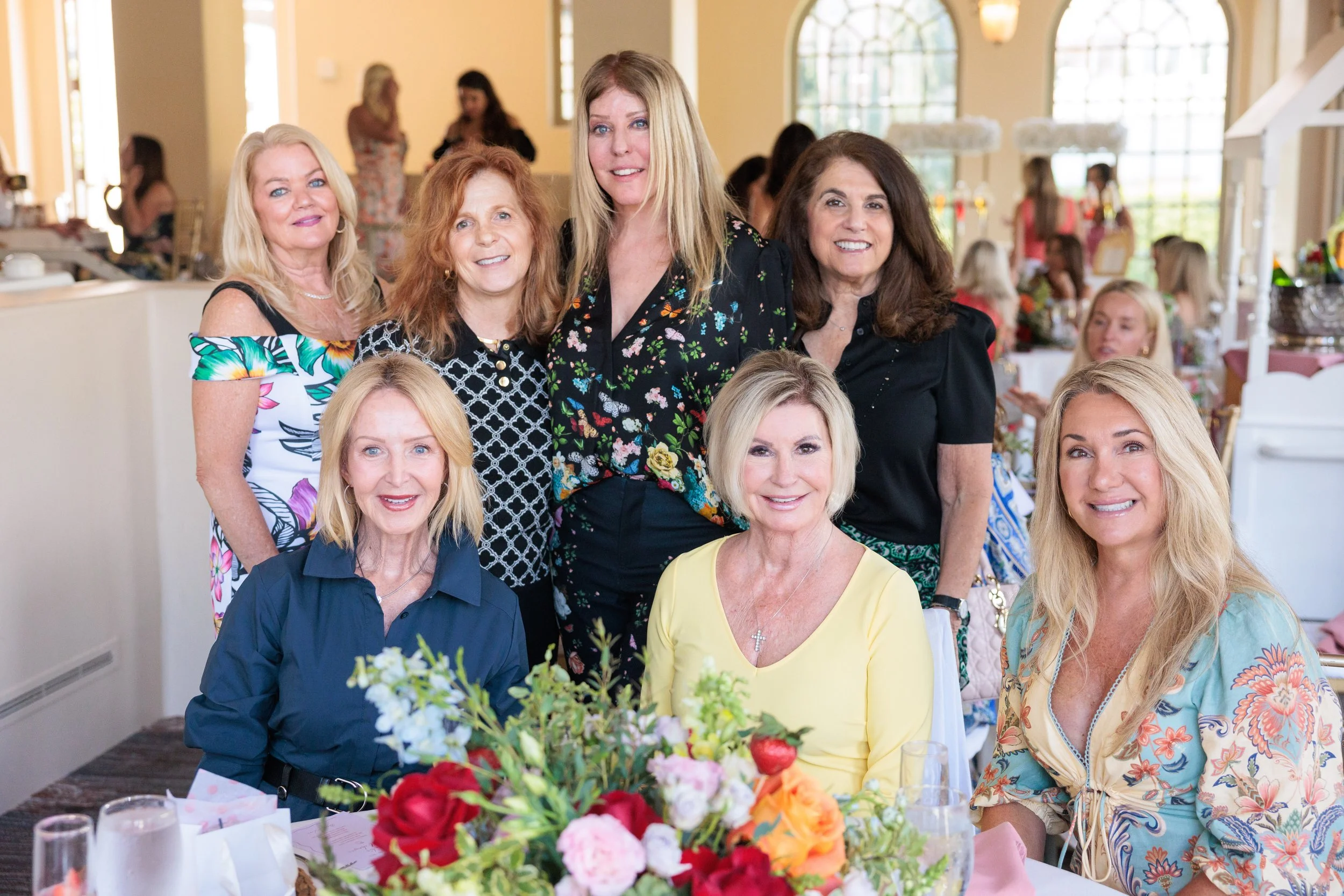 Group of nine women gathered at a celebration with tables decorated with flowers in a bright, indoor venue.