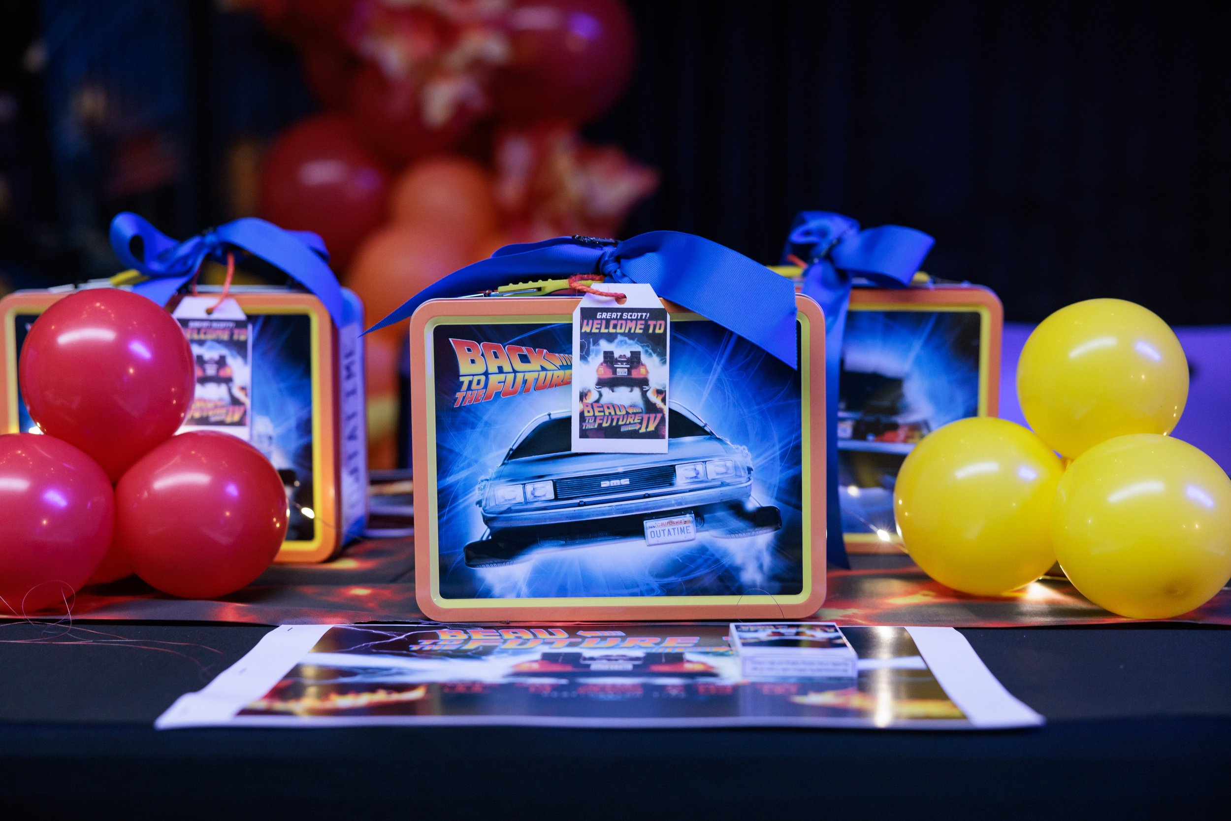 A display featuring 'Back to the Future' themed items, including a tin box with a hovercar on it, decorated with a blue ribbon, surrounded by pink and yellow balloons.