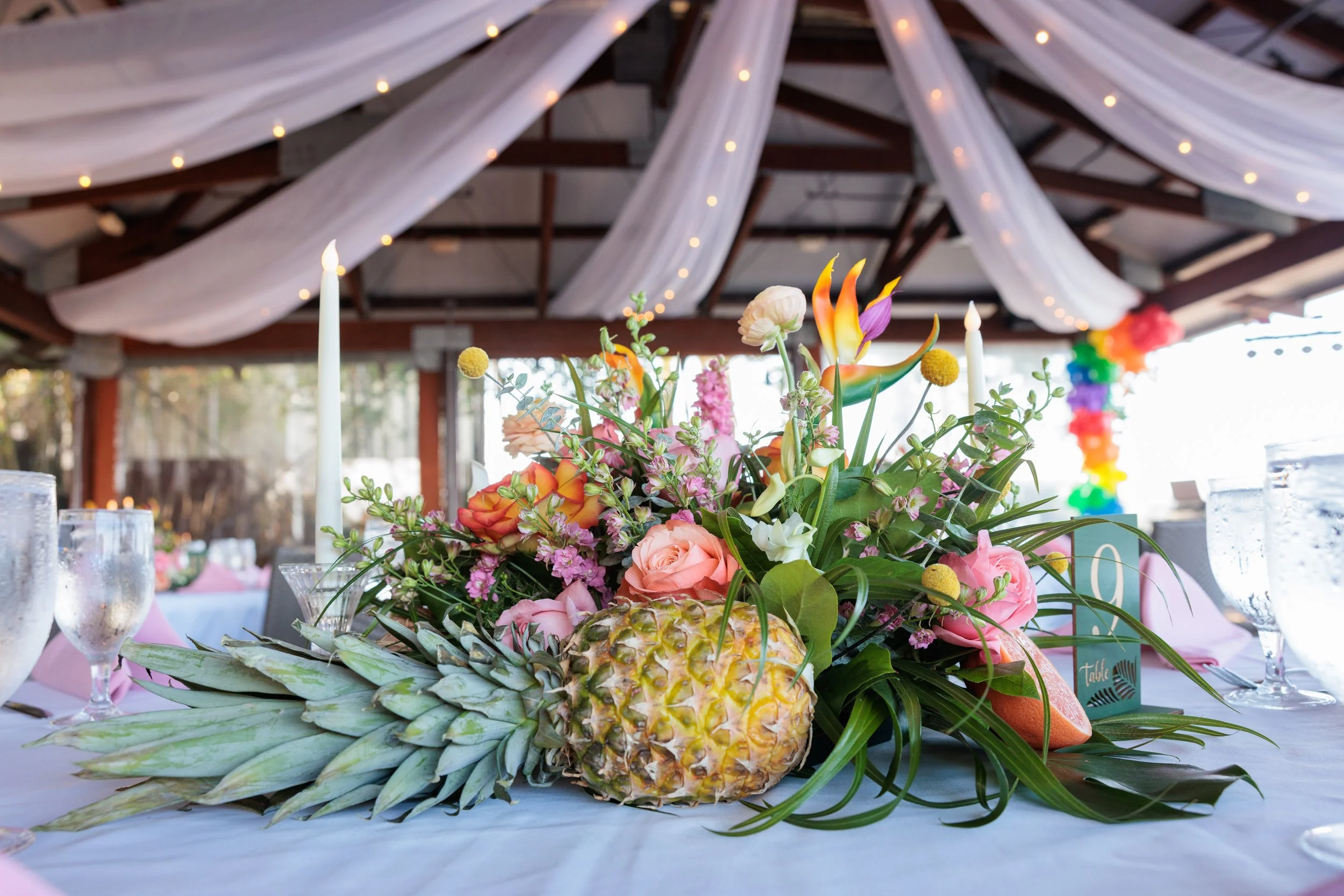 A floral centerpiece with pineapple, pink roses, orange and purple flowers, and greenery on a table at a decorated event venue.