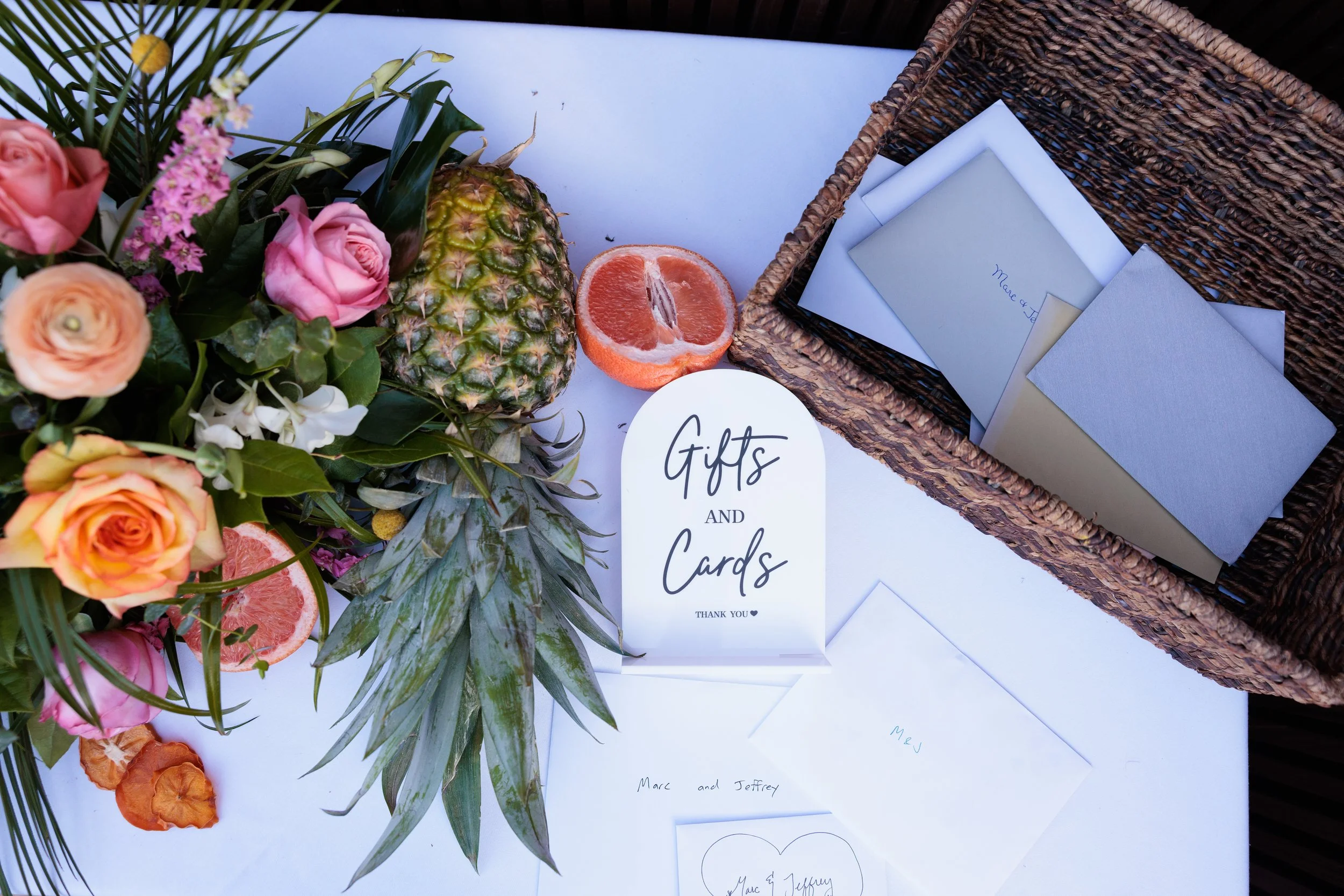 A display of gifts and cards, including a bouquet of pink, peach, and white flowers, a pineapple, a sliced grapefruit, a white card with 'Gifts and Cards' written on it, and several greeting cards and envelopes in a wicker basket.