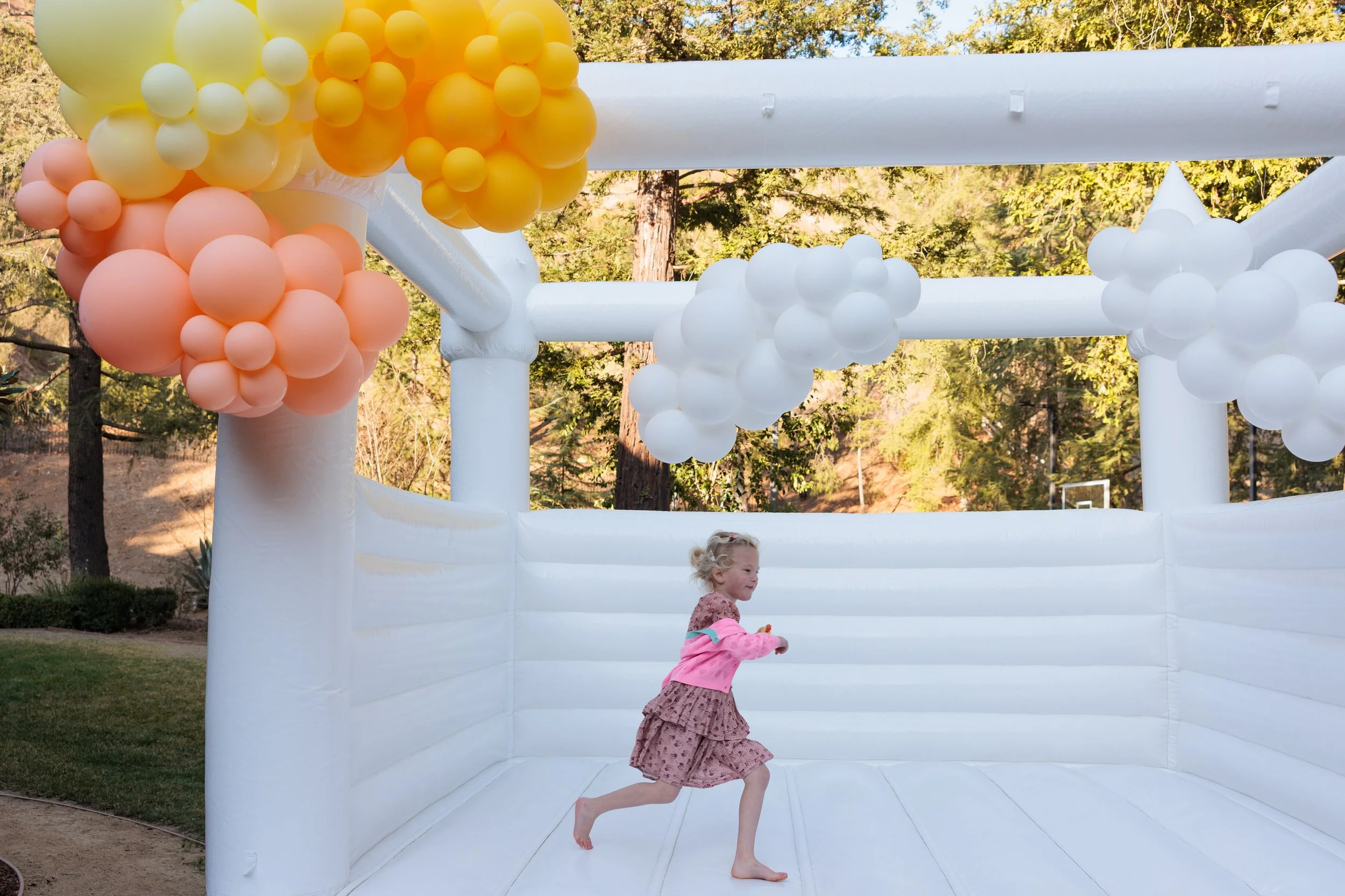 Young girl in a pink dress and hoodie playing inside a white inflatable bounce house decorated with peach, yellow, and white balloons, outdoors with green trees in the background.