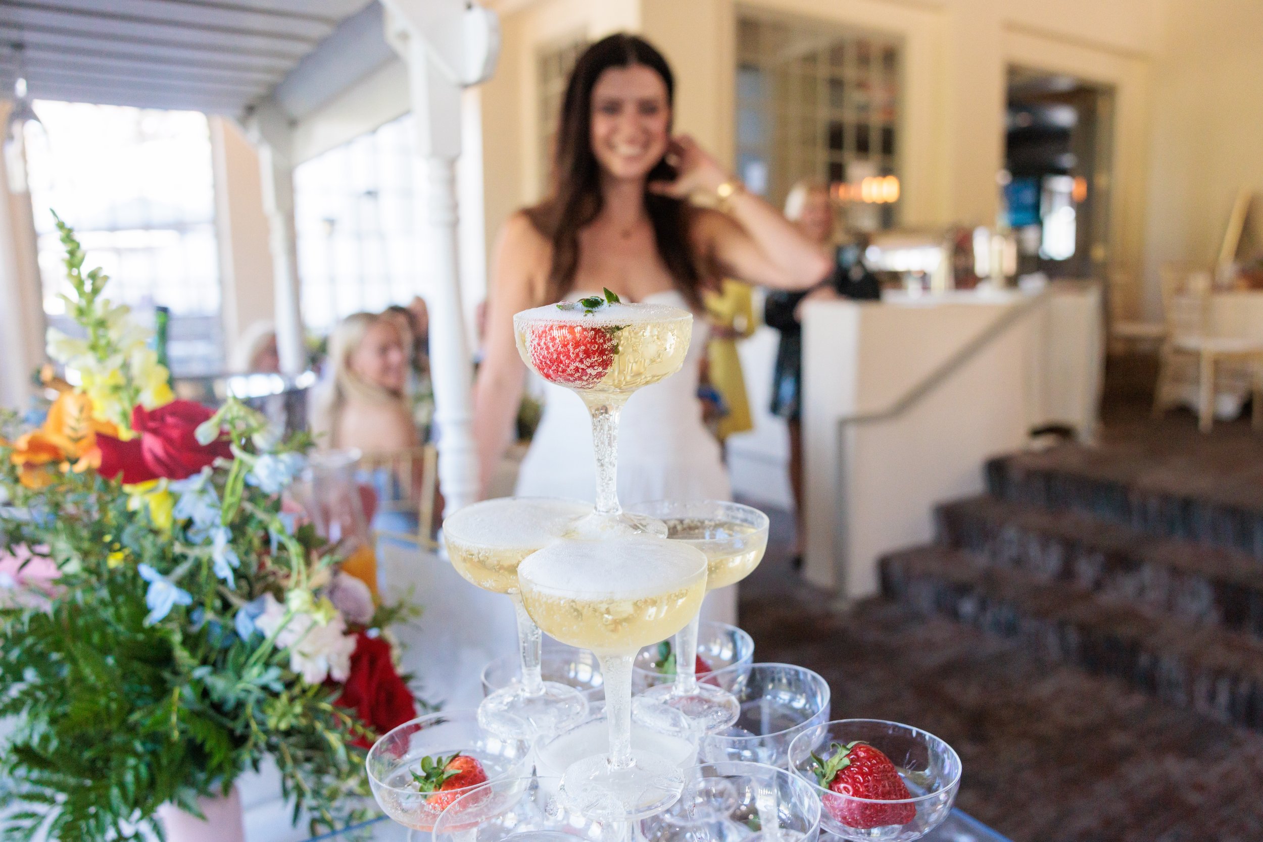 A tiered display of champagne glasses with champagne and strawberries, and a woman smiling in the background at a celebration event.