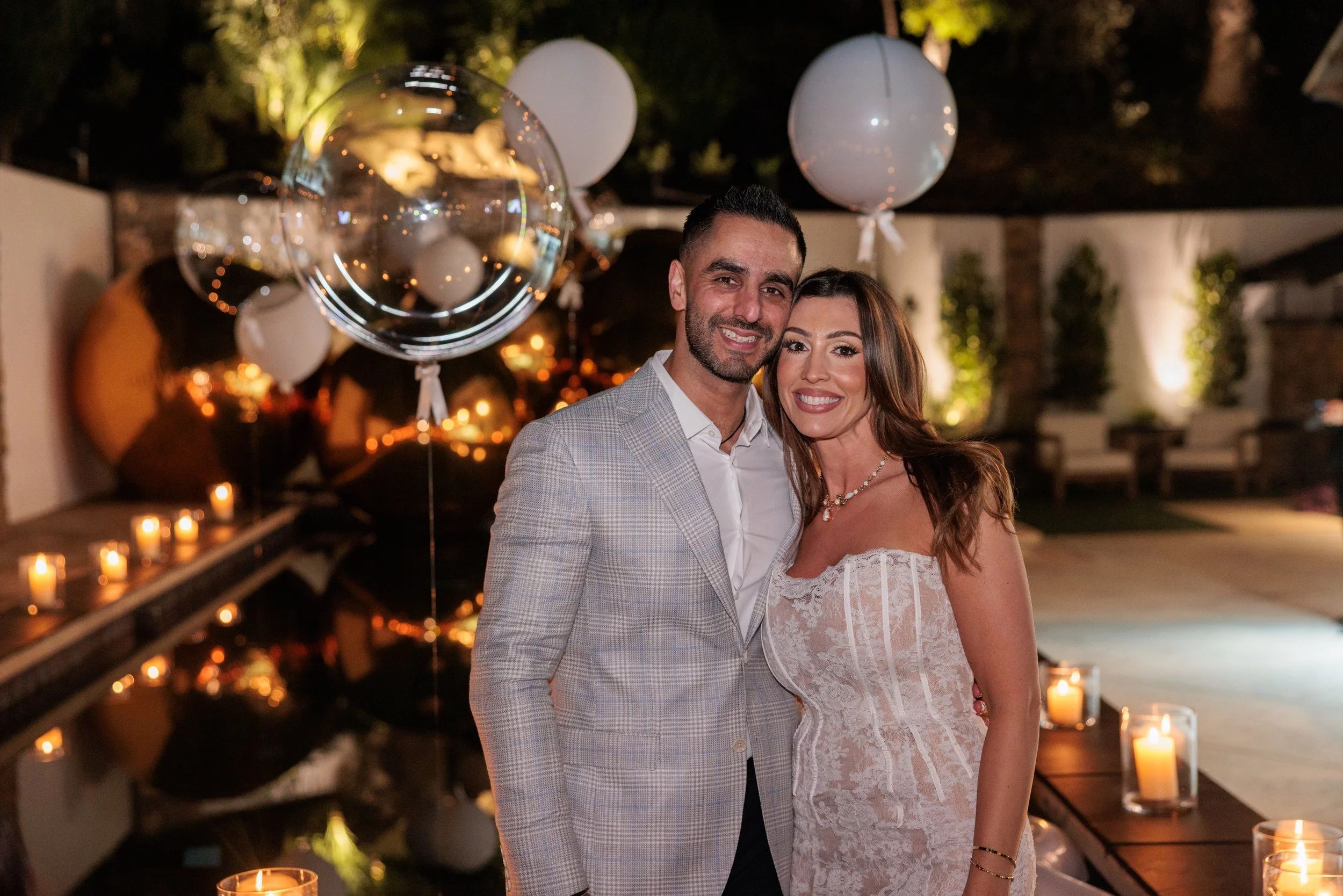 A happy couple dressed in formal attire, standing close together at an outdoor evening event, surrounded by floating balloons, candles, and warm decorative lighting.