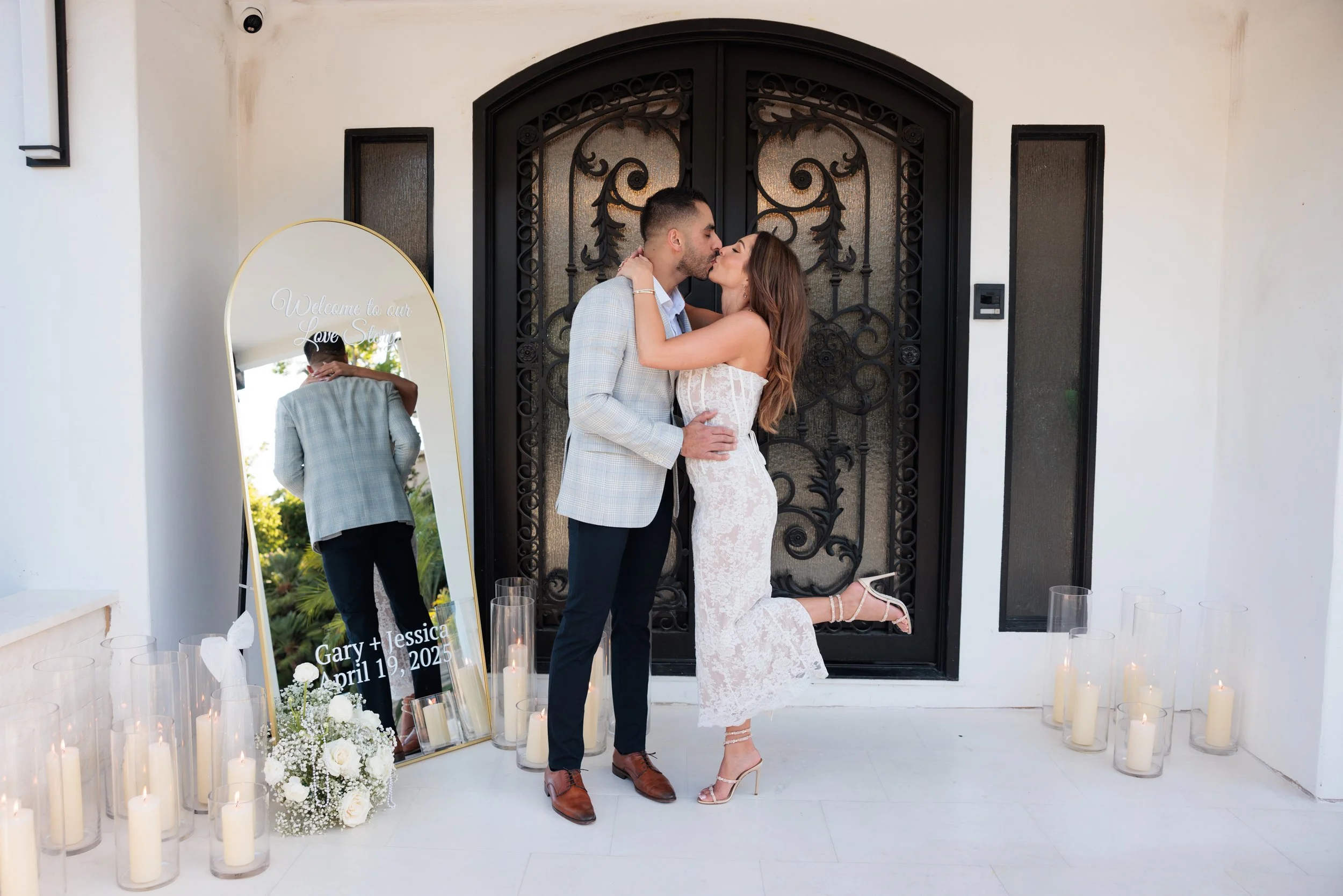 A couple sharing a kiss in front of a decorative black iron gate, with a large mirror reflecting the man, candles on the floor, and a floral arrangement, after their wedding. The mirror reads "Welcome to our Love Story," and has the names "Gary + Jes