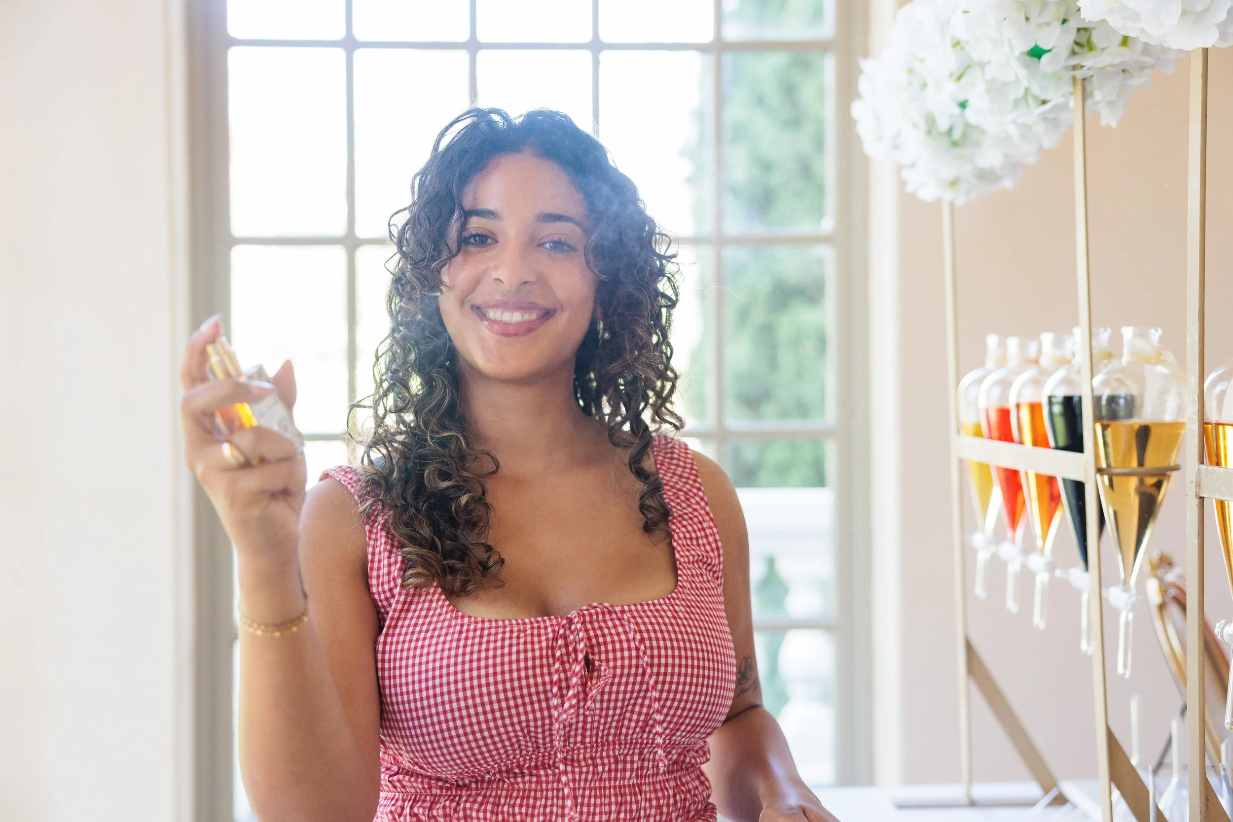 A smiling woman with curly hair in a red checkered dress holding a small bottle of perfume, standing inside a well-lit room with large window behind her and colorful liquids on the right.