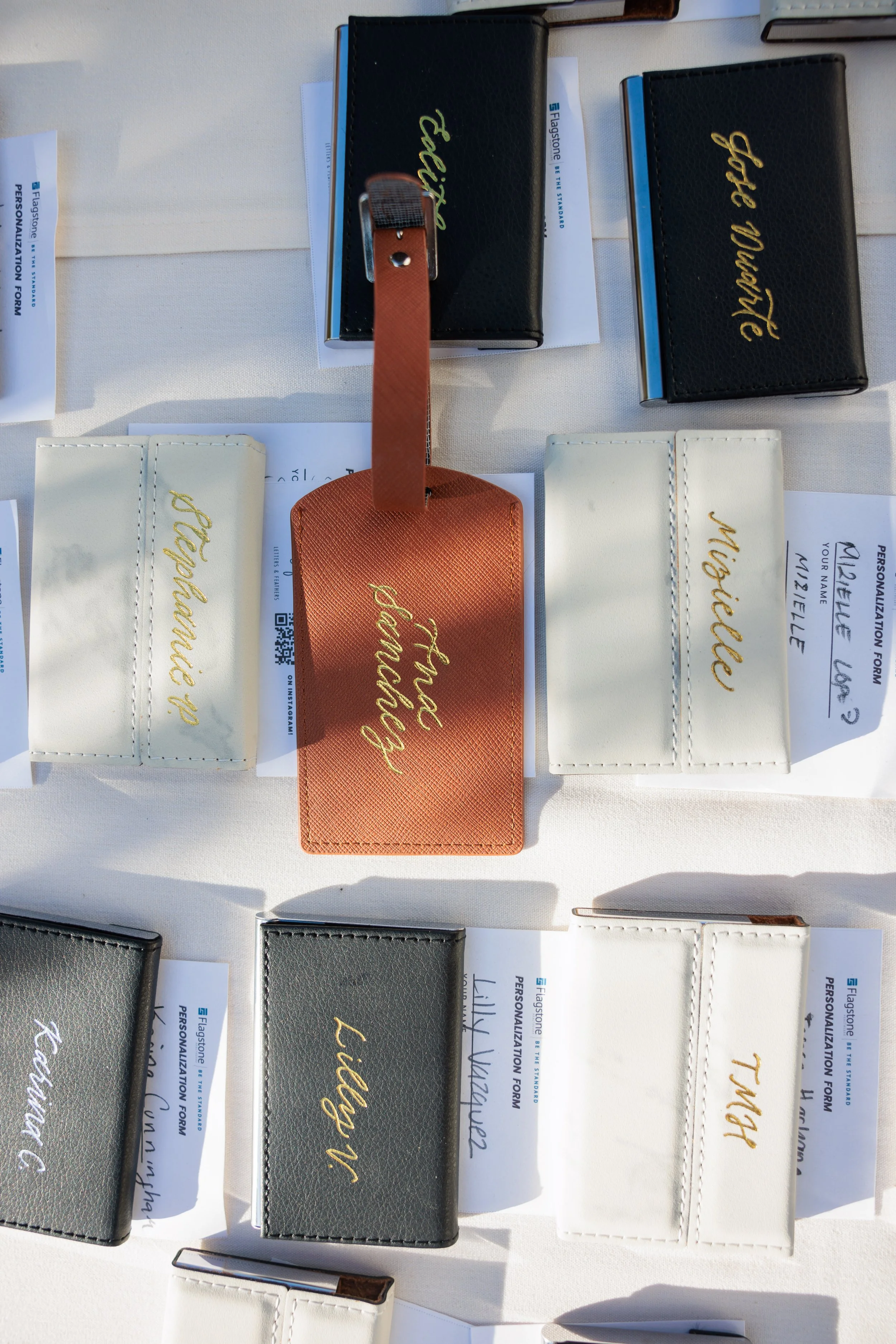 A display of various leather wallets on a table, each with a different name embossed in gold lettering. There are black, white, gray, and copper-colored wallets arranged neatly.