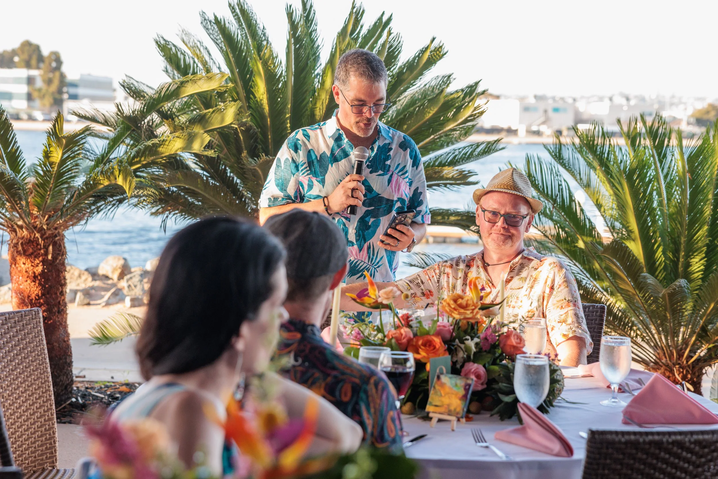 A man in a colorful Hawaiian shirt speaking into a microphone at an outdoor party or celebration, seated at a table with a floral centerpiece, surrounded by guests, with palm trees, water, and buildings in the background.