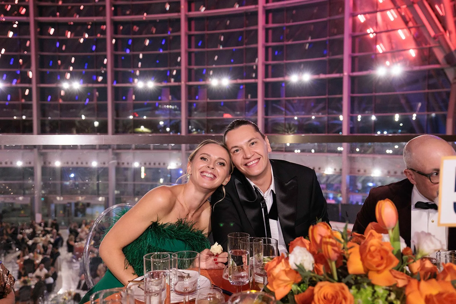 A smiling couple sitting at a banquet table during a formal event, with a man wearing a tuxedo and a woman in a green dress, in a large glass-covered venue with multicolored lights.
