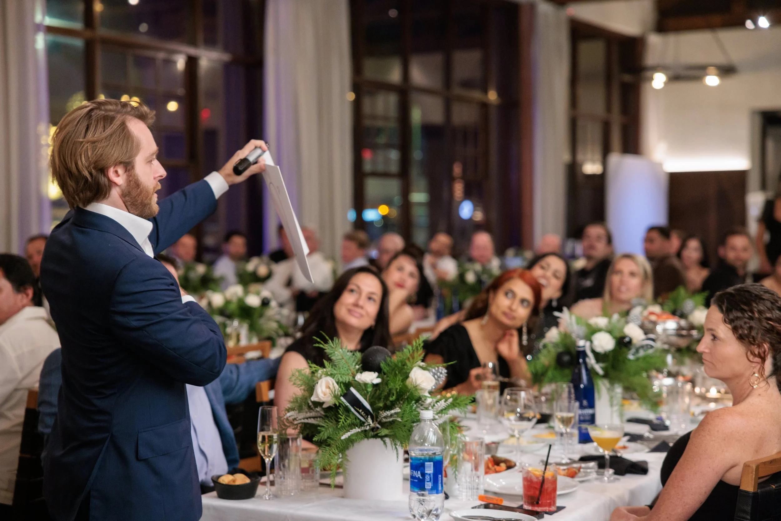 A man in a navy suit giving a speech at a banquet with guests seated at a decorated dining table.