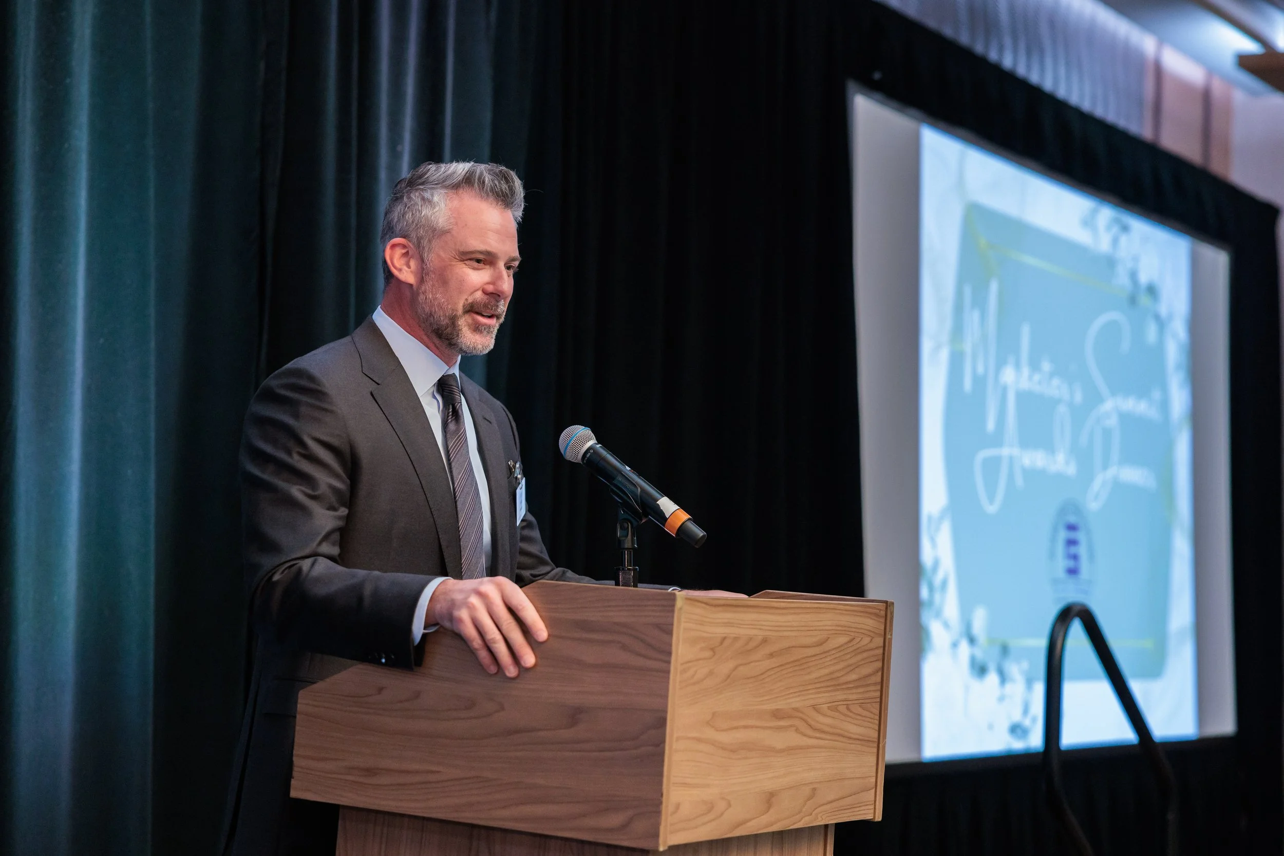 A middle-aged man with gray hair and a beard, wearing a dark suit, tie, and white shirt, stands at a wooden podium speaking into a microphone during a presentation or conference. Behind him is a large screen displaying a blue and white graphic.