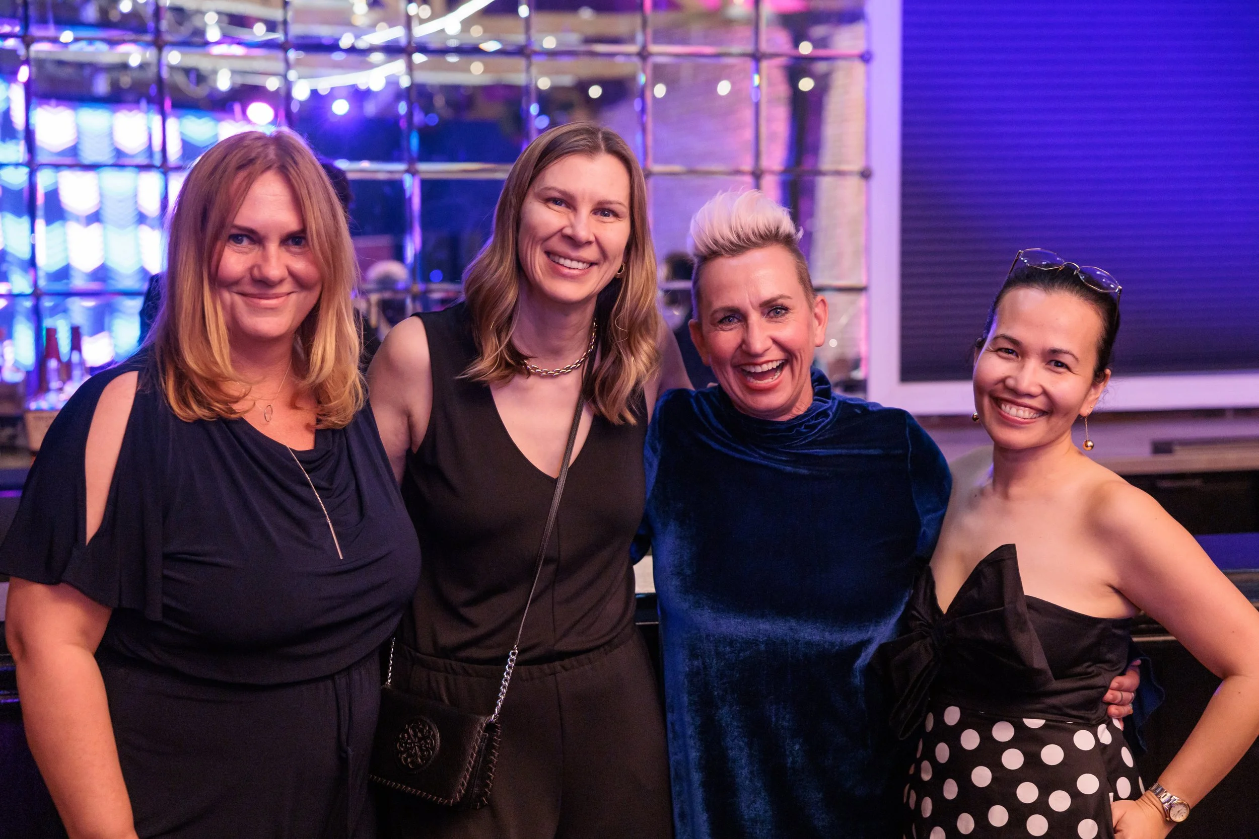 Four women smiling and posing together indoors at a social event with colorful lighting and a reflective background.