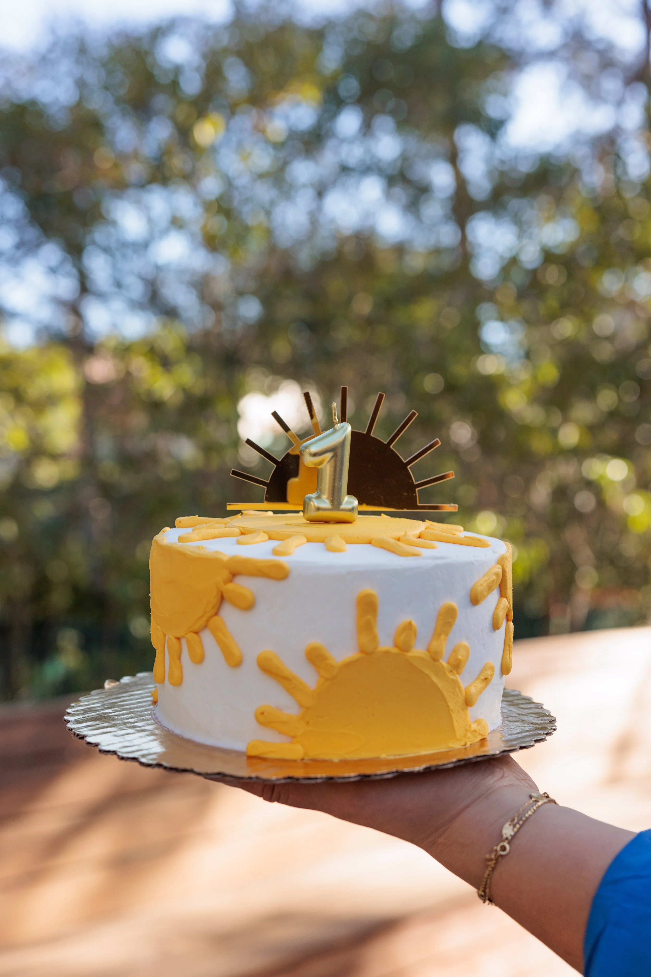 A birthday cake decorated with sun and sunrise motifs, topped with a number one candle and a sunburst decoration. The cake is held outdoors against a blurred background of trees and sky.