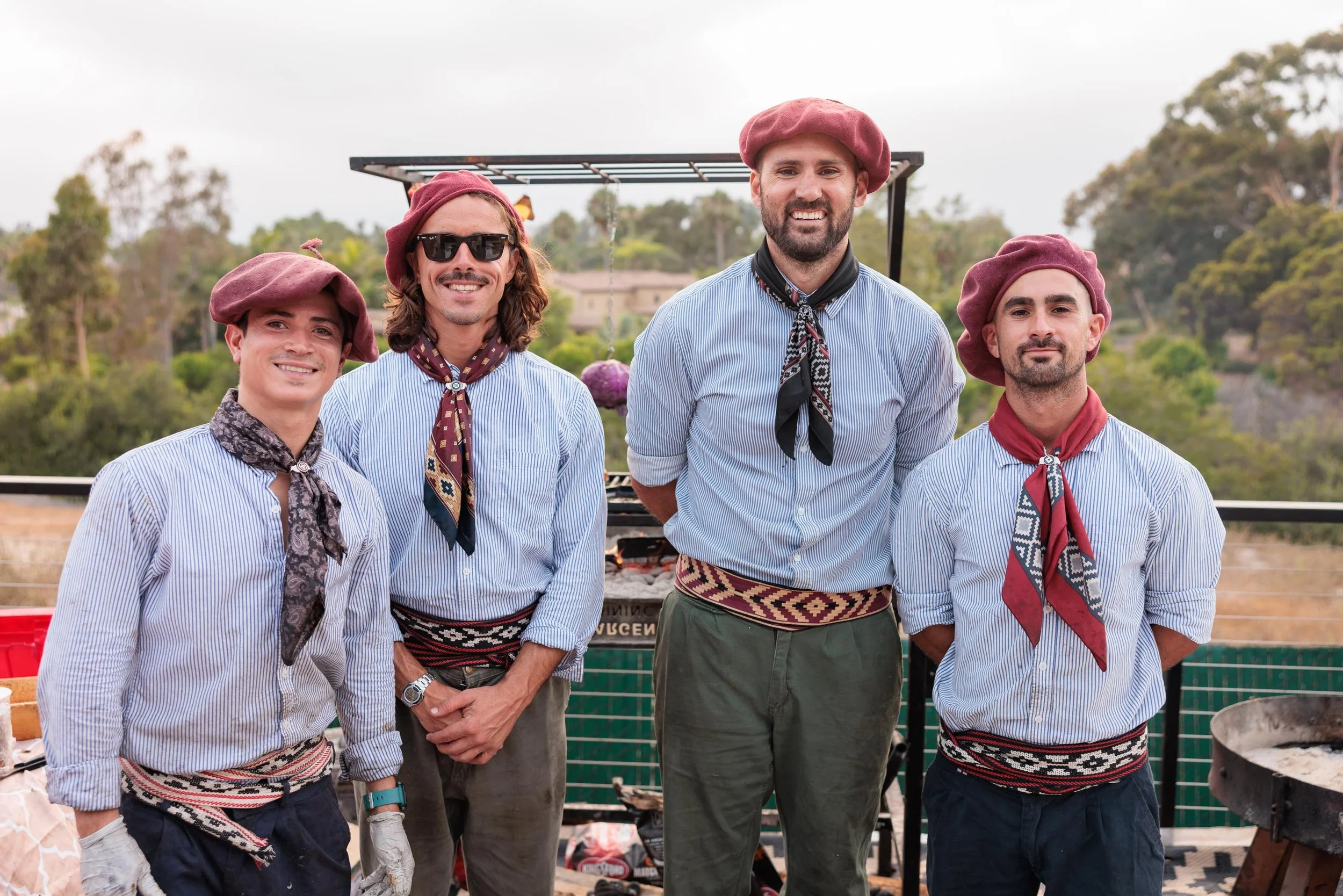 Four men dressed in blue and white striped shirts with traditional patterned sashes and maroon berets, standing outdoors with trees in the background, smiling at the camera.