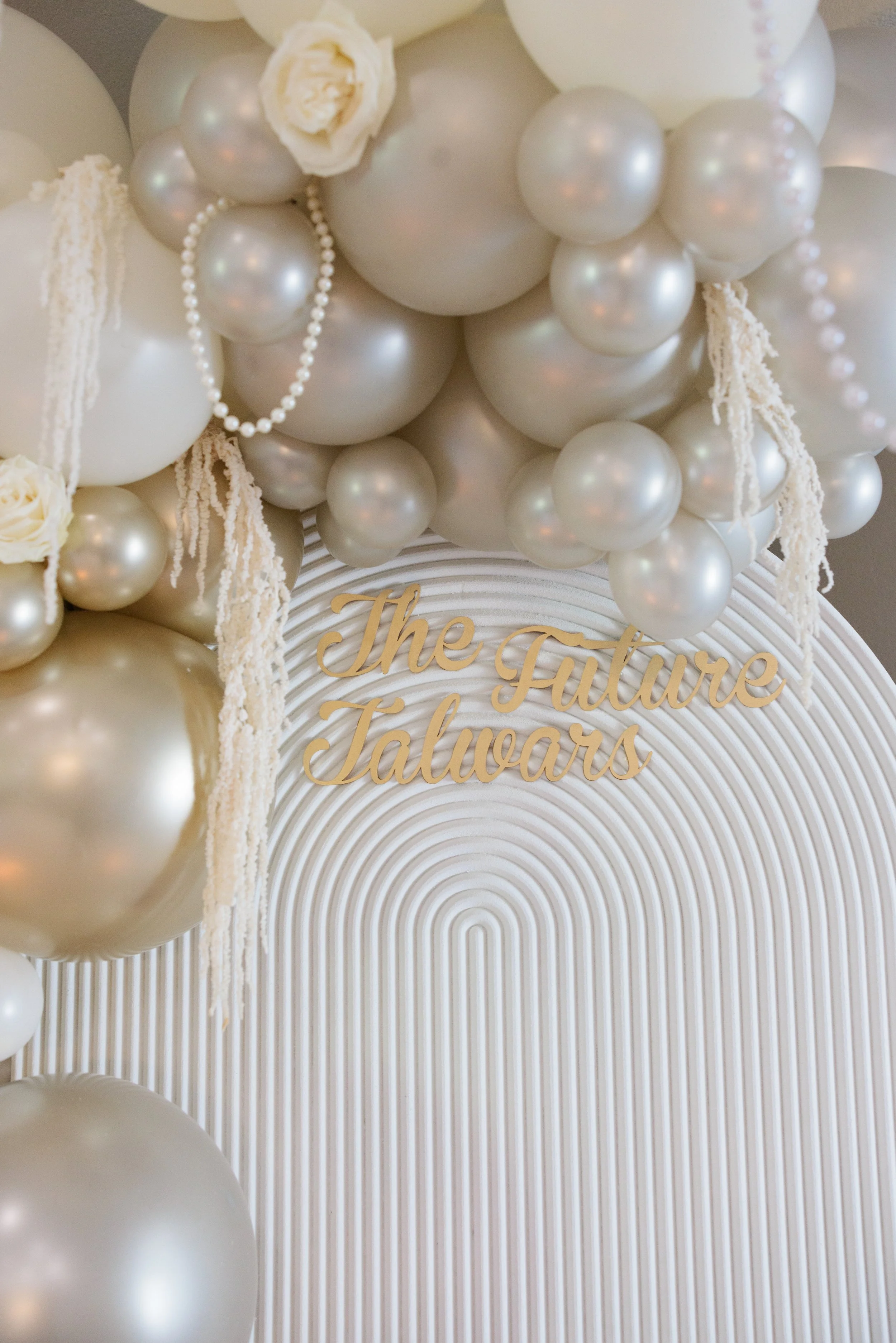 Decorative arrangement of white and pearl-colored balloons with white flowers and strings of pearls. A white, textured backdrop features gold cursive text reading 'The Future Talwars'.