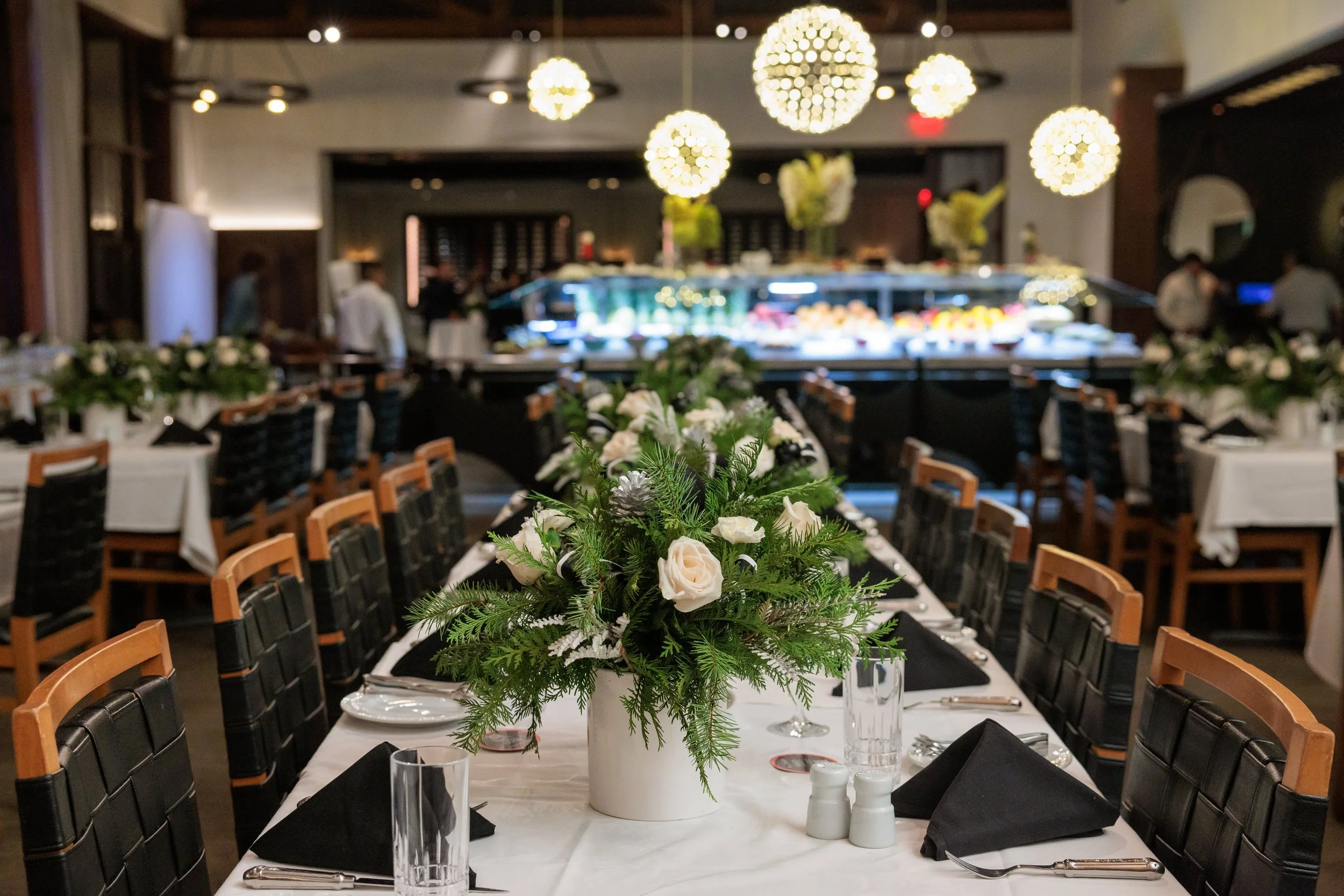 A beautifully set banquet table with a floral centerpiece featuring white roses and greenery in a white vase. The table is arranged with black napkins, glasses, and silverware, in an elegant restaurant or event space with decorative hanging lights an
