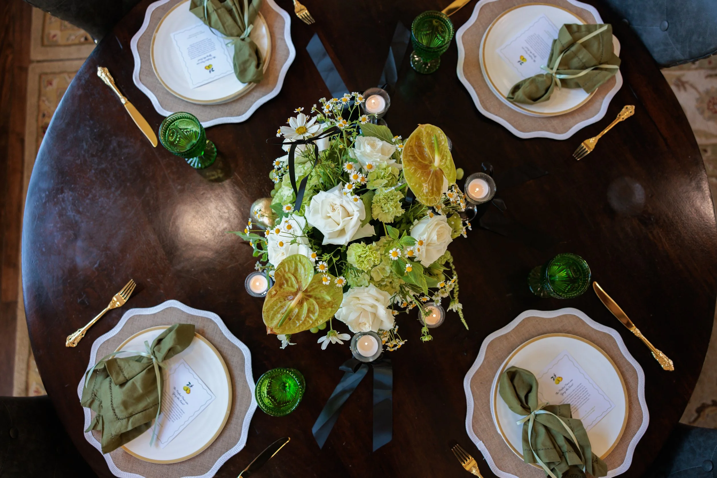 A round dining table set for four people with green glasses, gold utensils, white plates with beige and white placemats, green napkins tied with beige ribbon, and small candles. The center has a flower arrangement with white roses, daisies, and green