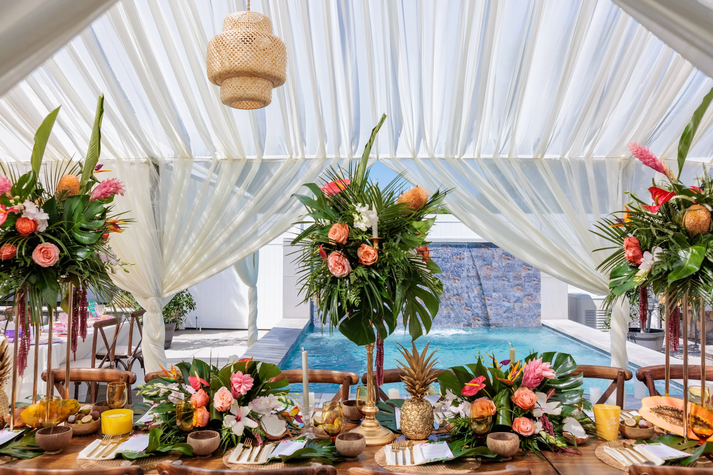 A decorated tropical-themed indoor event space with a table set for a meal, surrounded by floral arrangements and a view of a pool and waterfall through an open tent.