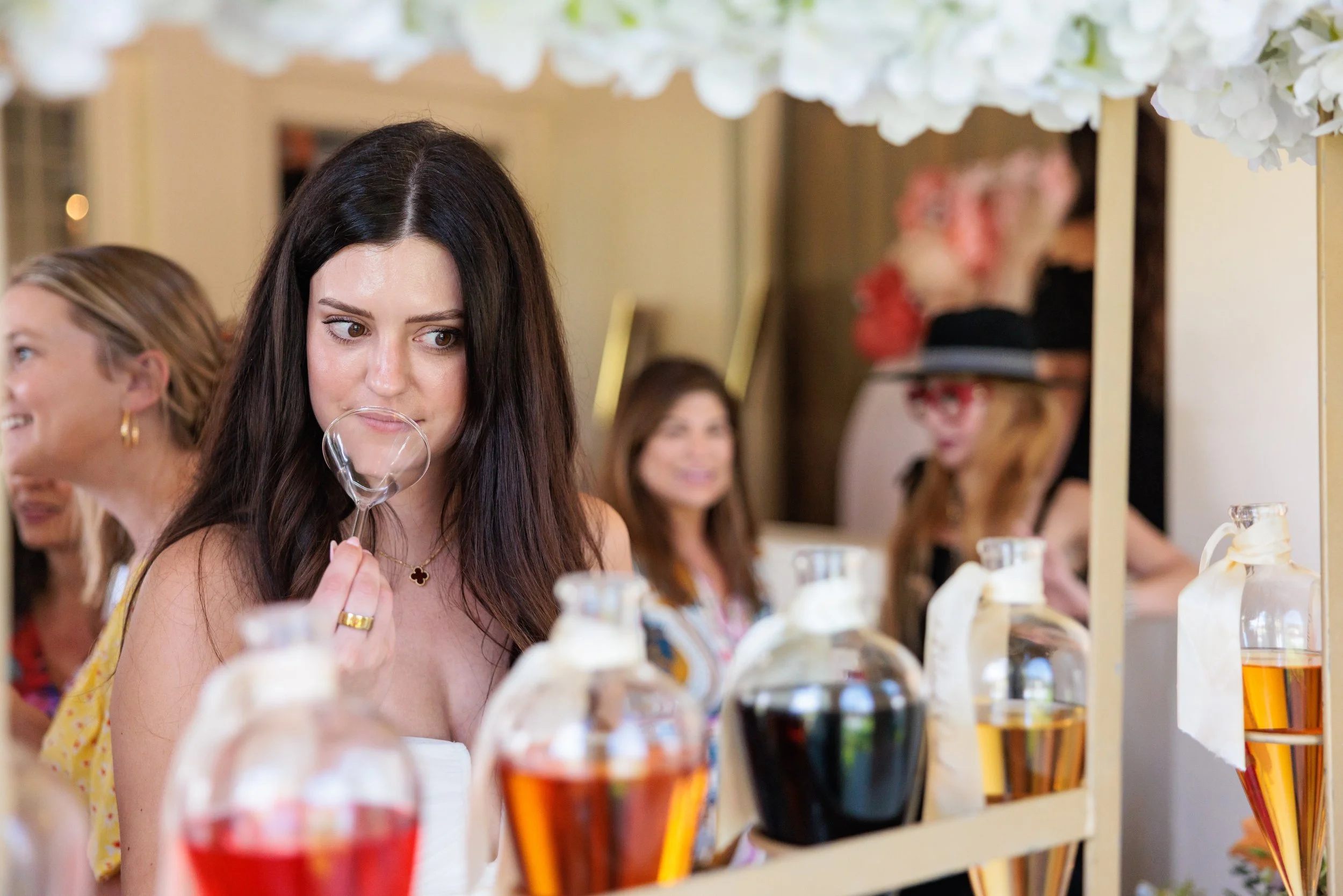 A woman with long dark hair, wearing a yellow dress, is smelling perfume at a display with various perfume bottles. She is in a well-lit indoor space, likely a boutique or event, with several women smiling and looking on in the background.