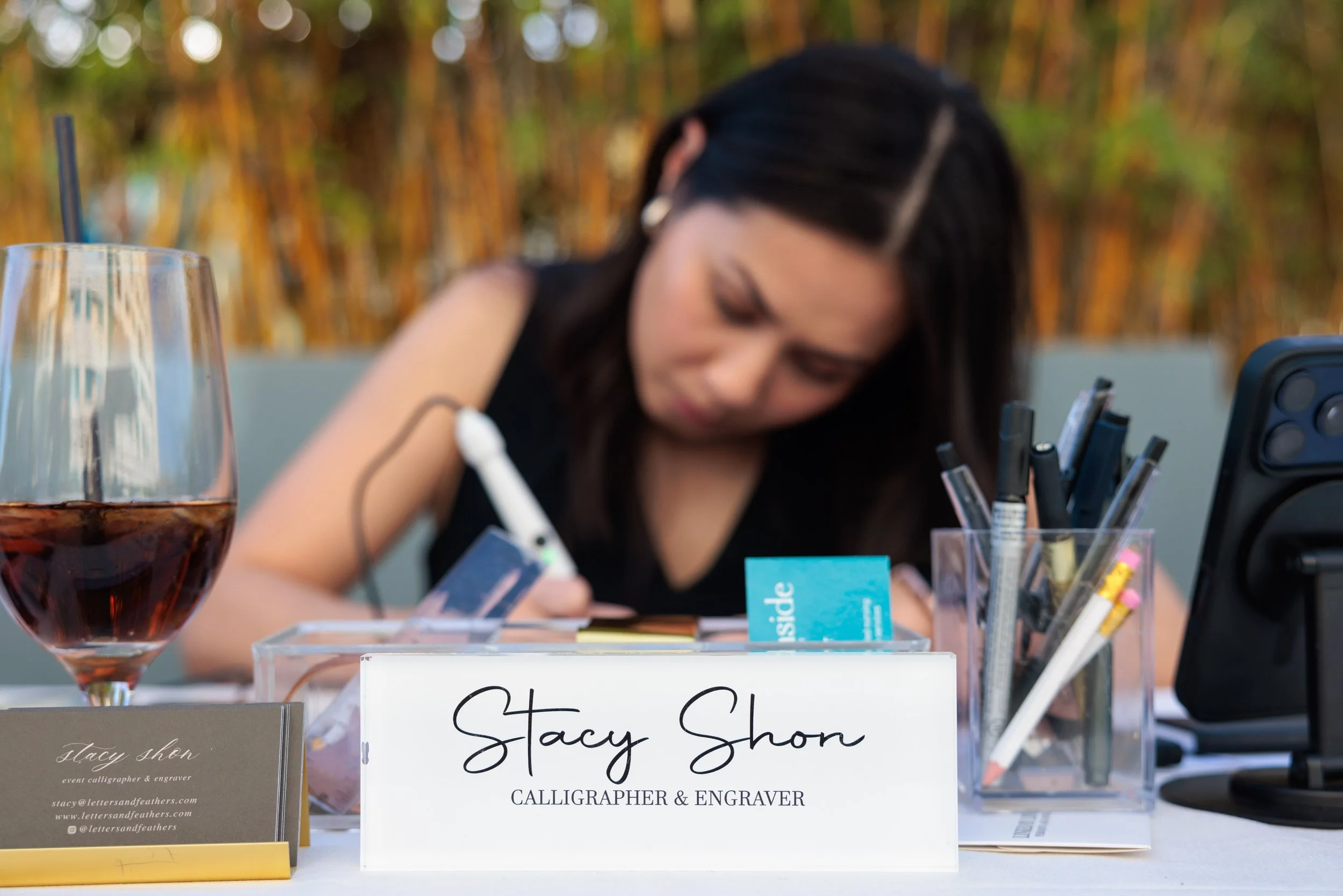 A woman sitting at a table writing with a pen, with a sign in front that reads 'Stacy Shon Calligrapher & Engraver', a glass of red wine, business cards, and containers holding pens and markers.