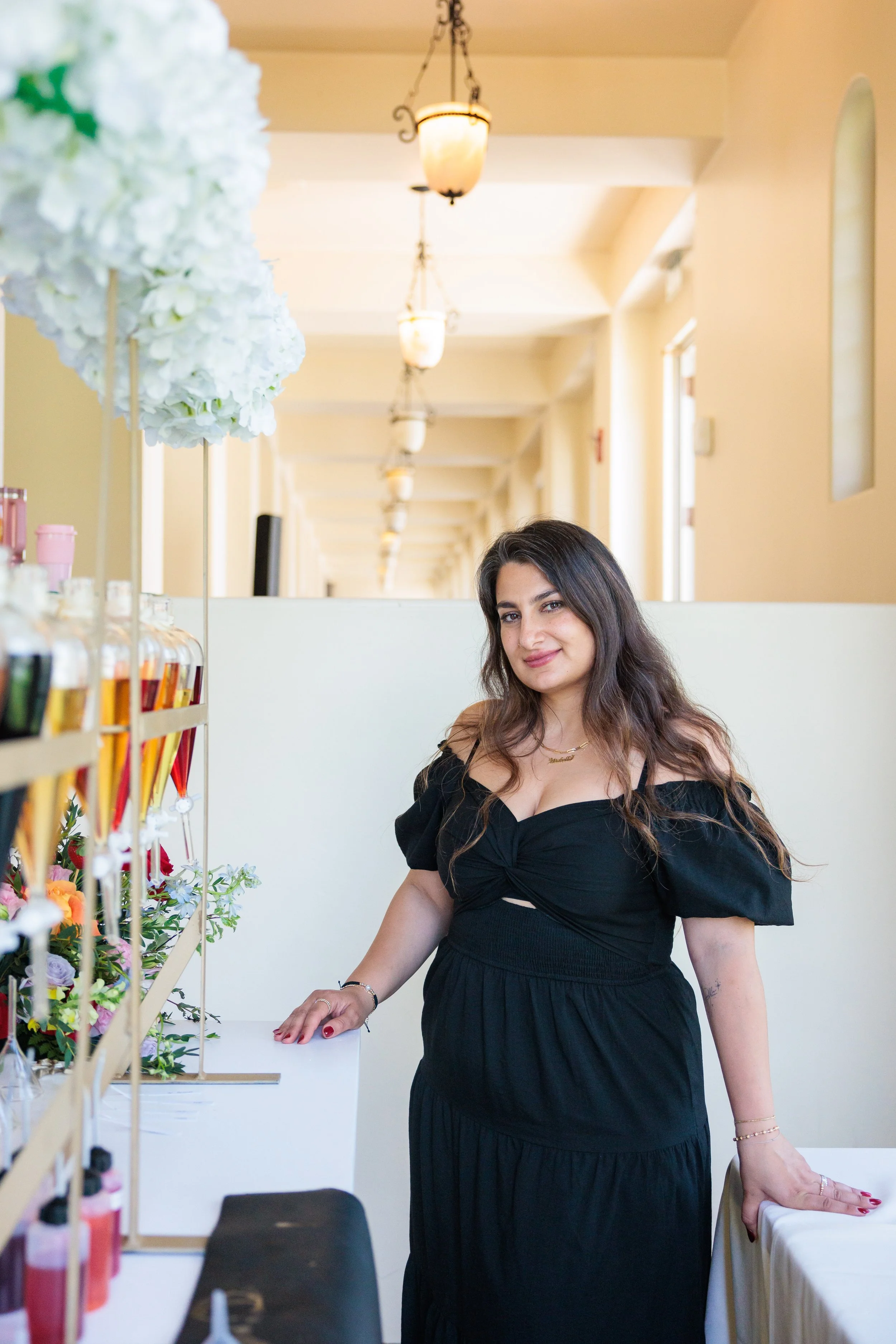 Young woman in a black dress standing near a display of colorful perfume bottles and flowers in an elegant indoor setting with hanging light fixtures.