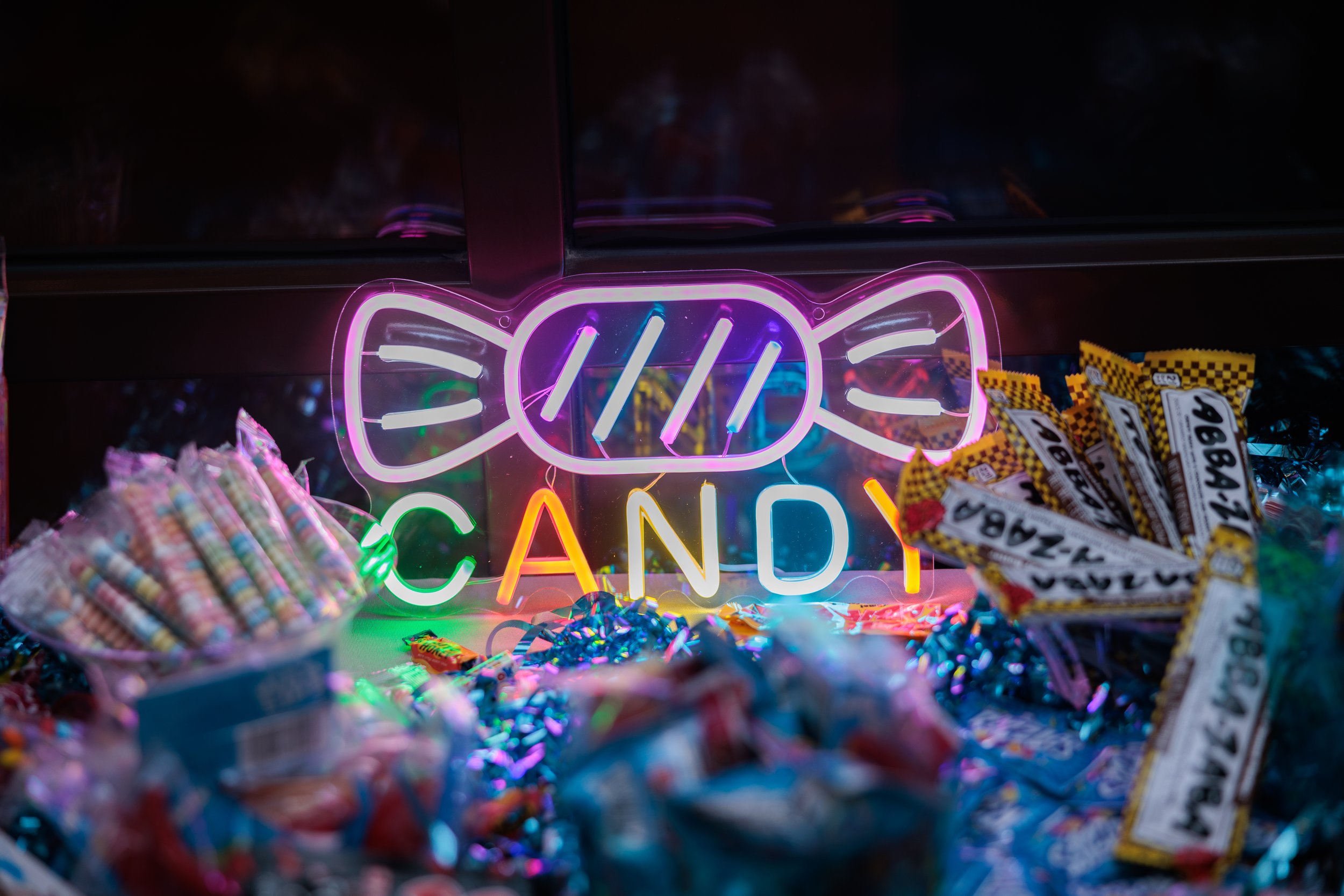 Neon sign reading "CANDY" with a wrapped candy illustration, surrounded by various candies in a display case.
