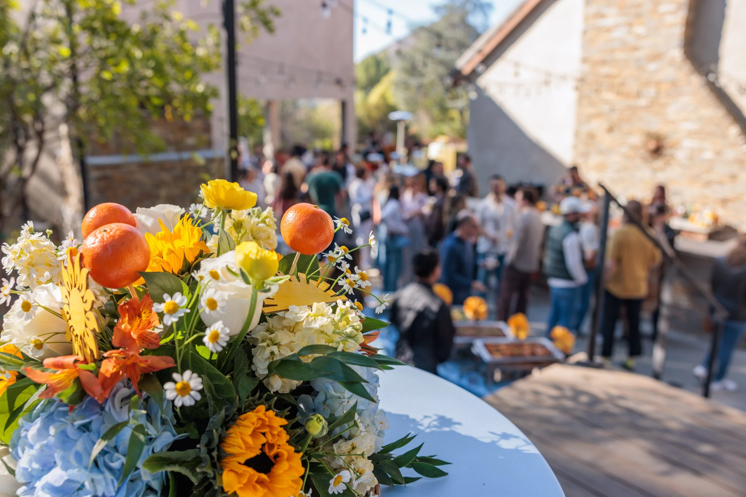 Close-up of a colorful flower arrangement with oranges, daisies, sunflowers, and other blooms, in the foreground at an outdoor gathering with people mingling in the background under string lights and in front of a stone and wooden wall.