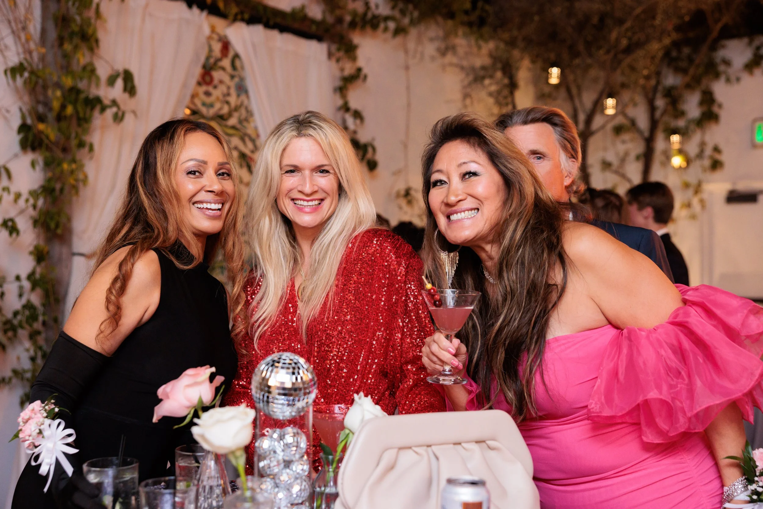 Three women smiling at a party, one holding a pink cocktail, with a decorated table and other guests in the background.