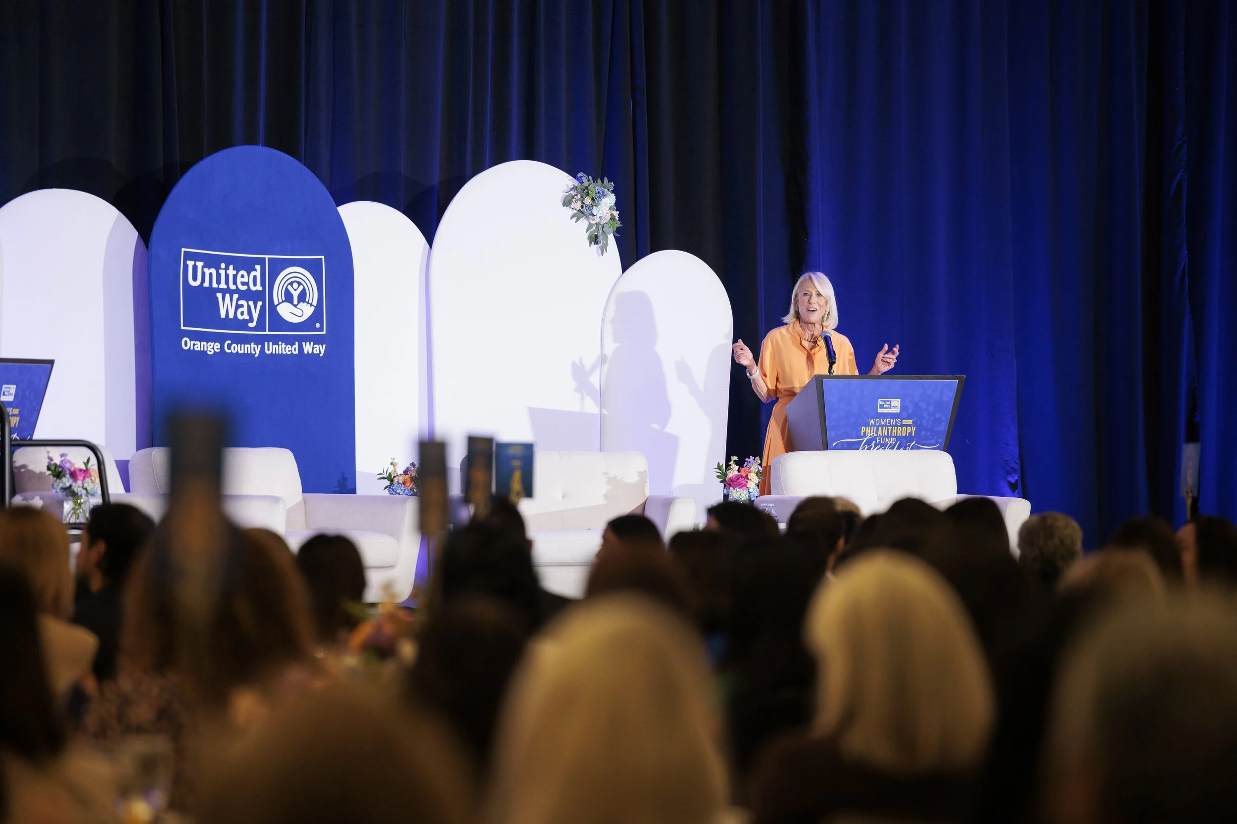 A woman speaking at a podium during a public event, with a large blue and white backdrop displaying the 'United Way' logo and 'Orange County United Way' above her on stage. The stage is decorated with flowers and white furniture, and there is an audi
