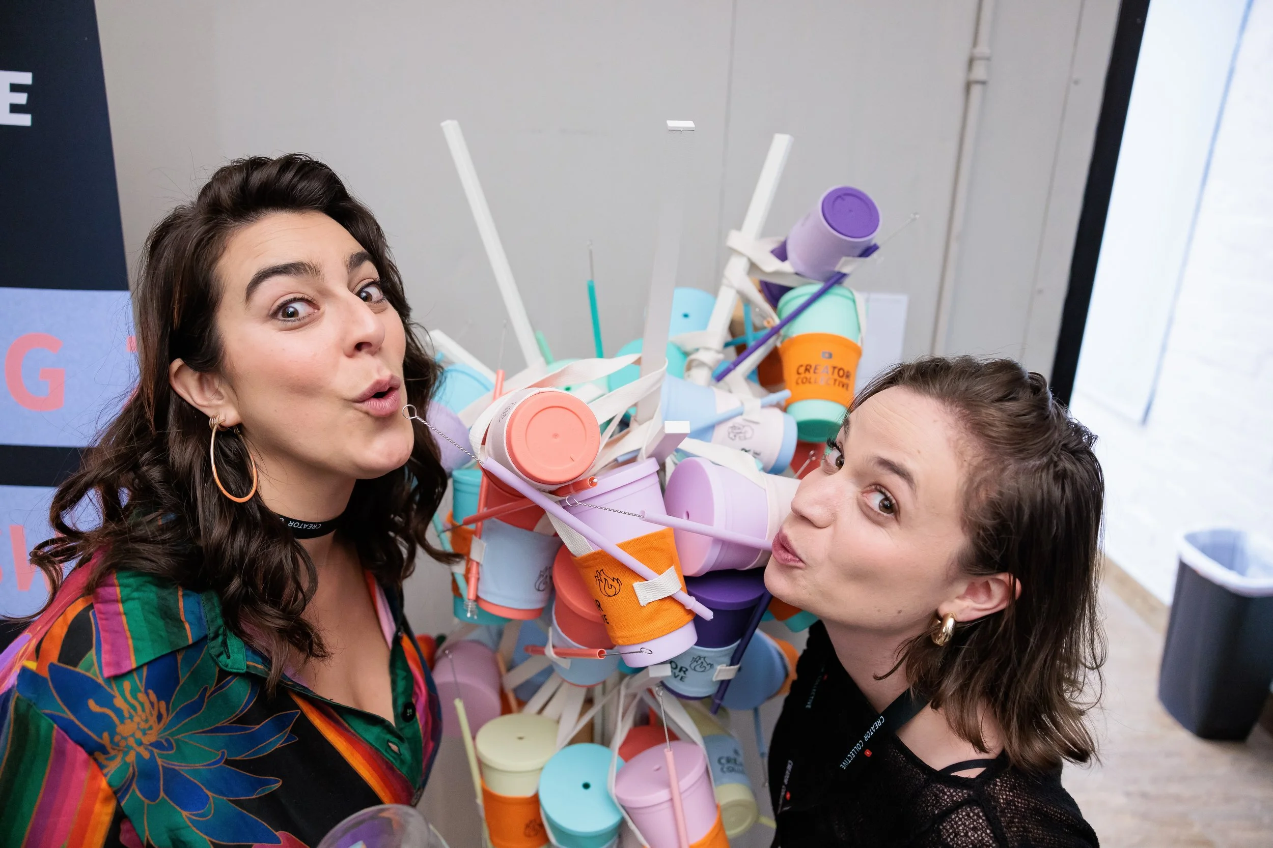Two women smiling and making playful faces next to a colorful pile of bubble tea cups with straws.